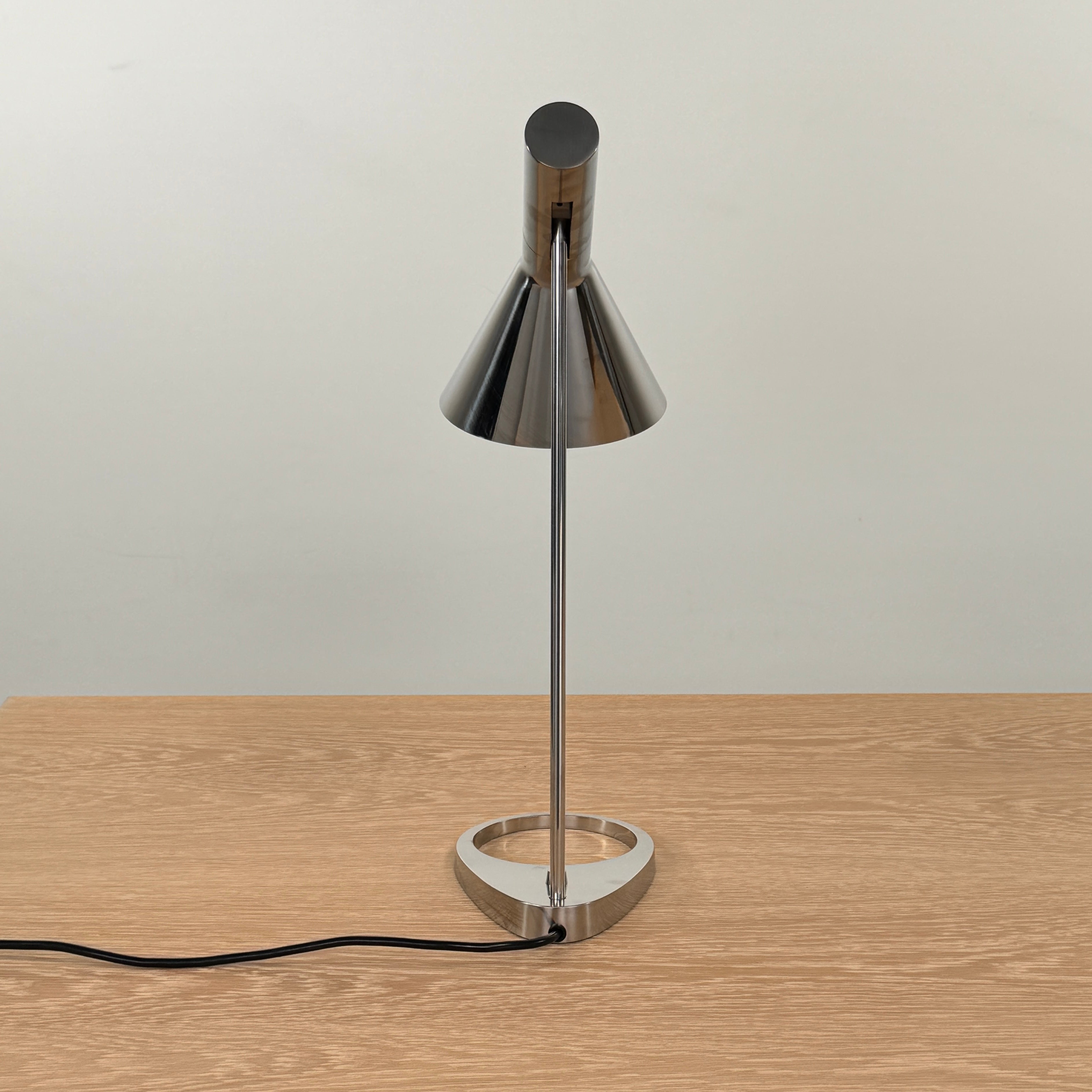 The Louis Poulsen AJ Table Lamp, featuring a modern metallic finish and slanted shade, rests on a wooden surface and illuminates the desk.