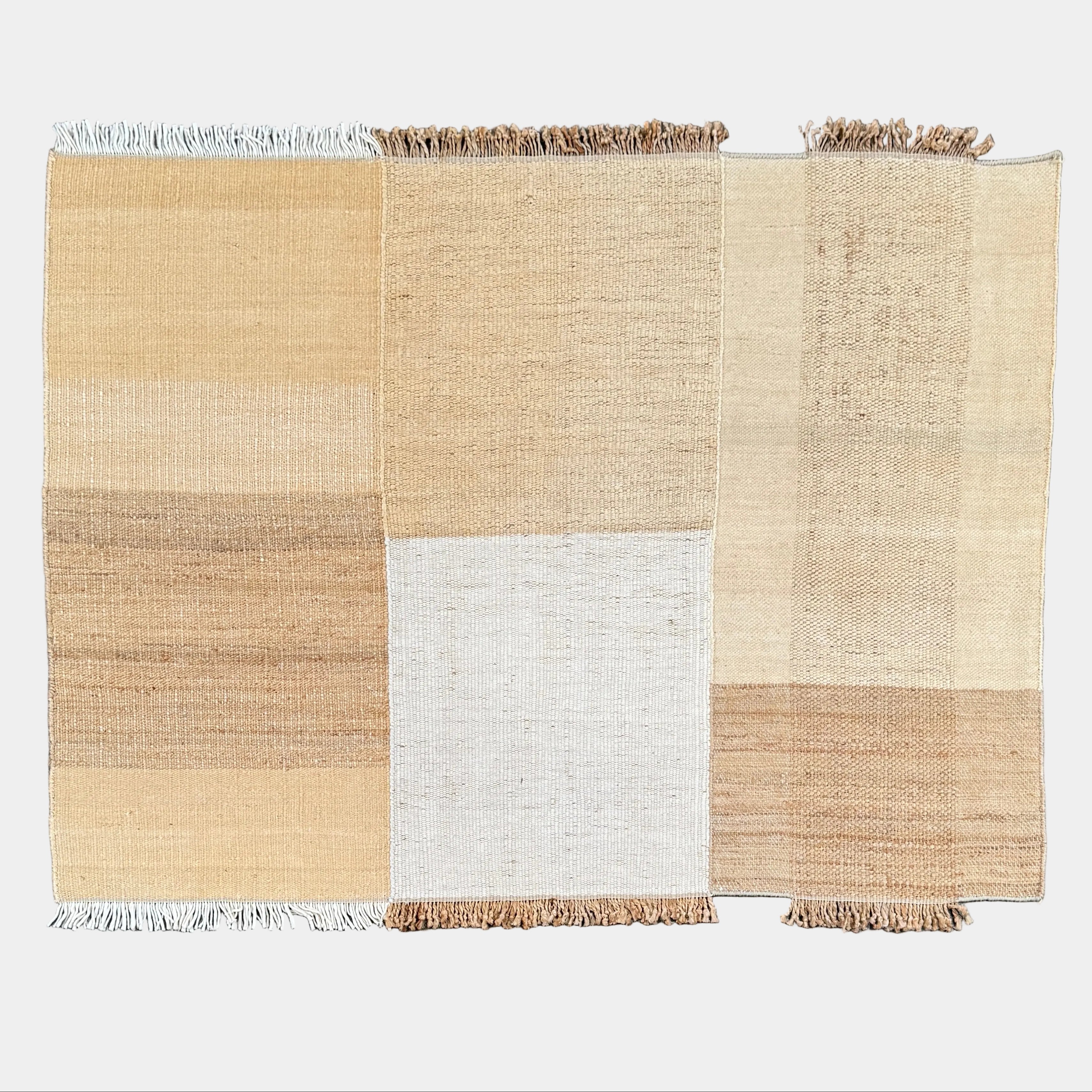 A rectangular Nanimarquina Tres Rug in Ocher, showcasing a patchwork design in warm beige and brown hues, is displayed on a concrete floor. This artisanal piece by Nanimarquina features hand-woven techniques and decorative fringe along two opposite edges, reminiscent of the brand’s signature style.