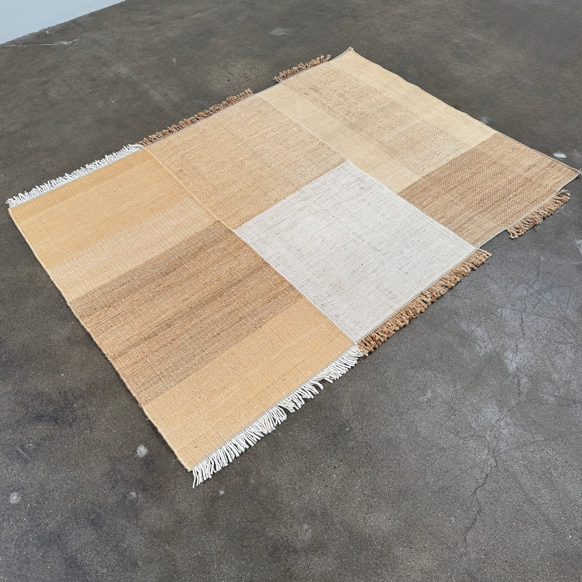 A rectangular Nanimarquina Tres Rug in Ocher, showcasing a patchwork design in warm beige and brown hues, is displayed on a concrete floor. This artisanal piece by Nanimarquina features hand-woven techniques and decorative fringe along two opposite edges, reminiscent of the brand’s signature style.