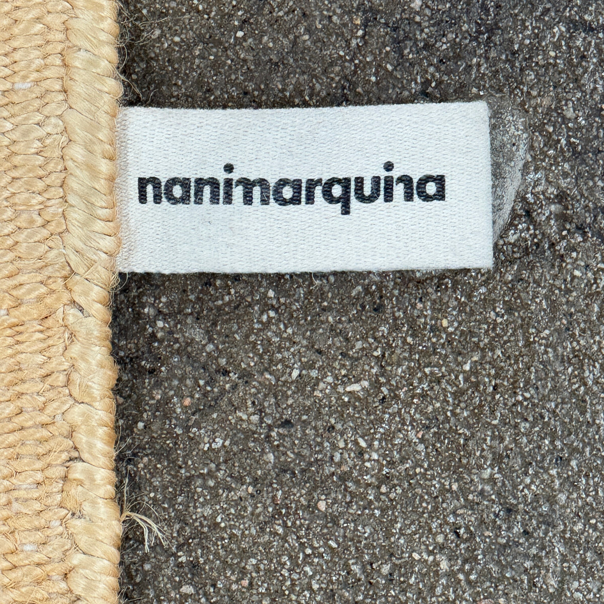 A rectangular Nanimarquina Tres Rug in Ocher, showcasing a patchwork design in warm beige and brown hues, is displayed on a concrete floor. This artisanal piece by Nanimarquina features hand-woven techniques and decorative fringe along two opposite edges, reminiscent of the brand’s signature style.