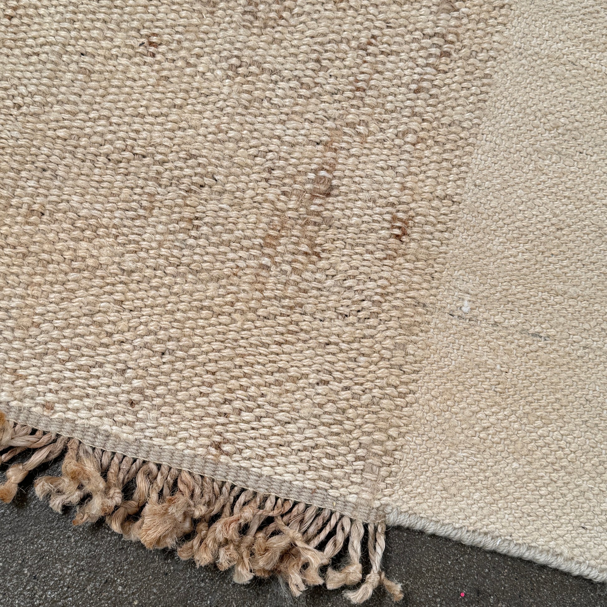 A rectangular Nanimarquina Tres Rug in Ocher, showcasing a patchwork design in warm beige and brown hues, is displayed on a concrete floor. This artisanal piece by Nanimarquina features hand-woven techniques and decorative fringe along two opposite edges, reminiscent of the brand’s signature style.