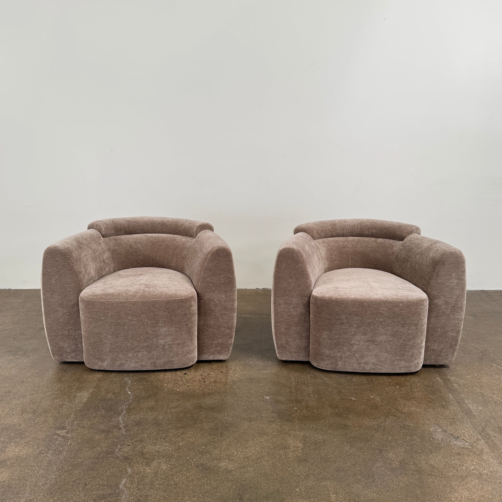 Two Daniel Boddam Wave Lounge Chairs in Maharam Mohair, featuring plush taupe upholstery with rounded backs and arms, are arranged side by side on a polished concrete floor against a plain white wall.