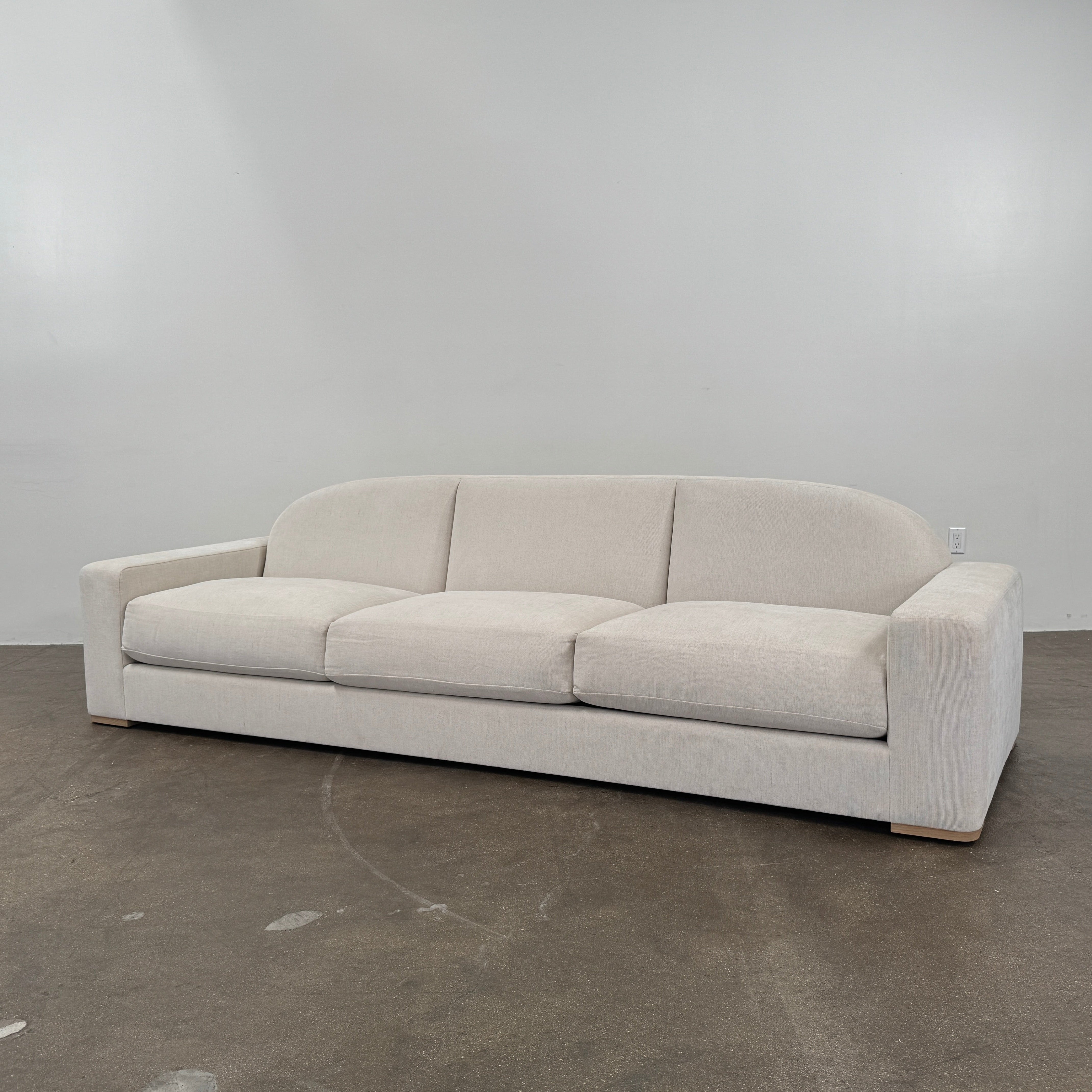 The Dmitriy & Co. Erato Sofa by Dimitriy & Co is a light gray three-seater featuring low arms and wooden feet, showcased against a plain white background—an elegant example of bespoke furnishings and couture upholstery.