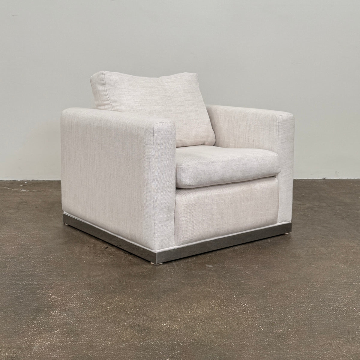 Two identical Milo Baughman-style armchairs from Vintage, in a light beige color with boxy shapes and cushions, are arranged side by side on a bare concrete floor against a plain light wall, forming an elegant “Milo Baughman - In the style of - Armchair Set of 2.”.