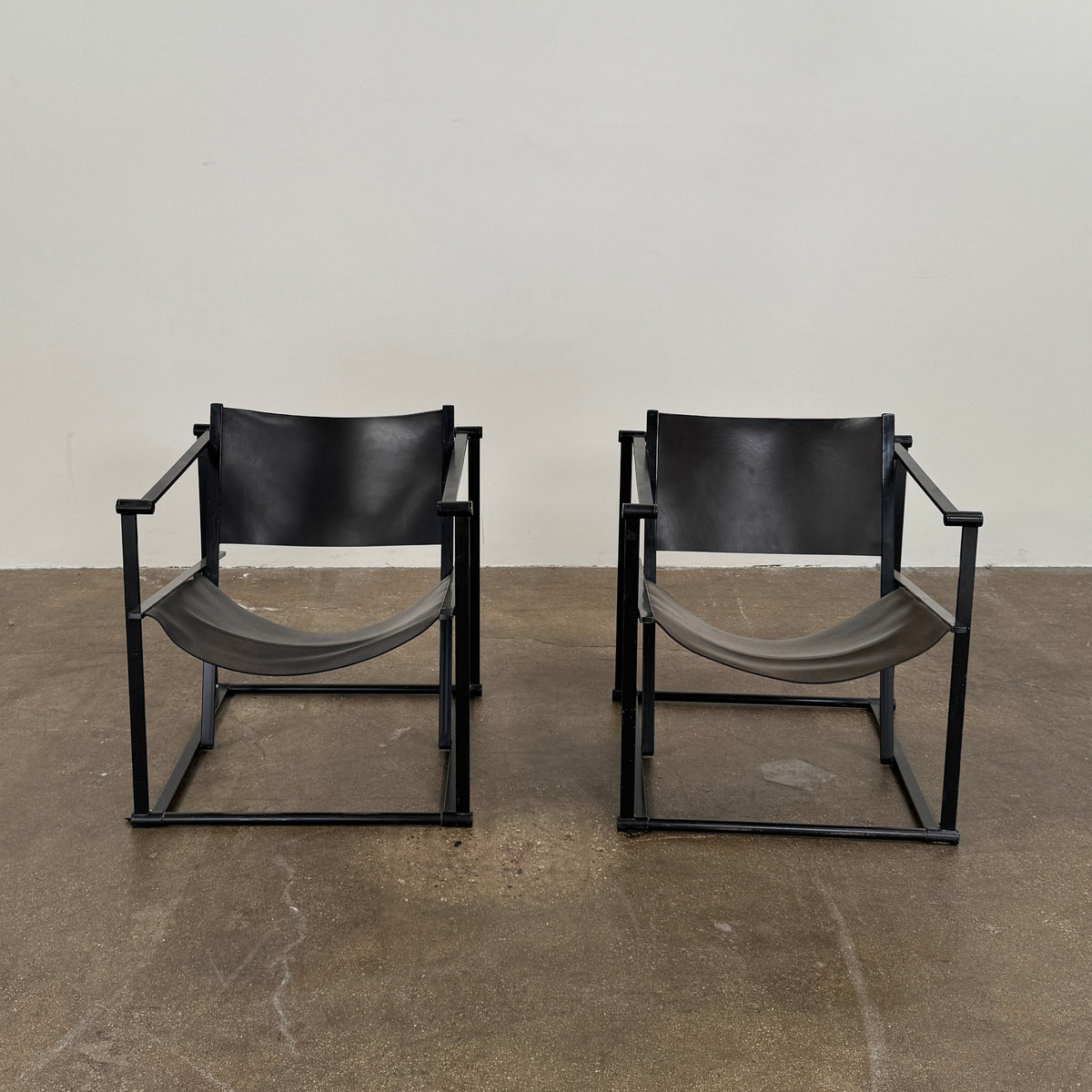 Two Pastoe FM62 chairs in black leather by Modern Resale are positioned side by side on a brown concrete floor against a plain white wall.