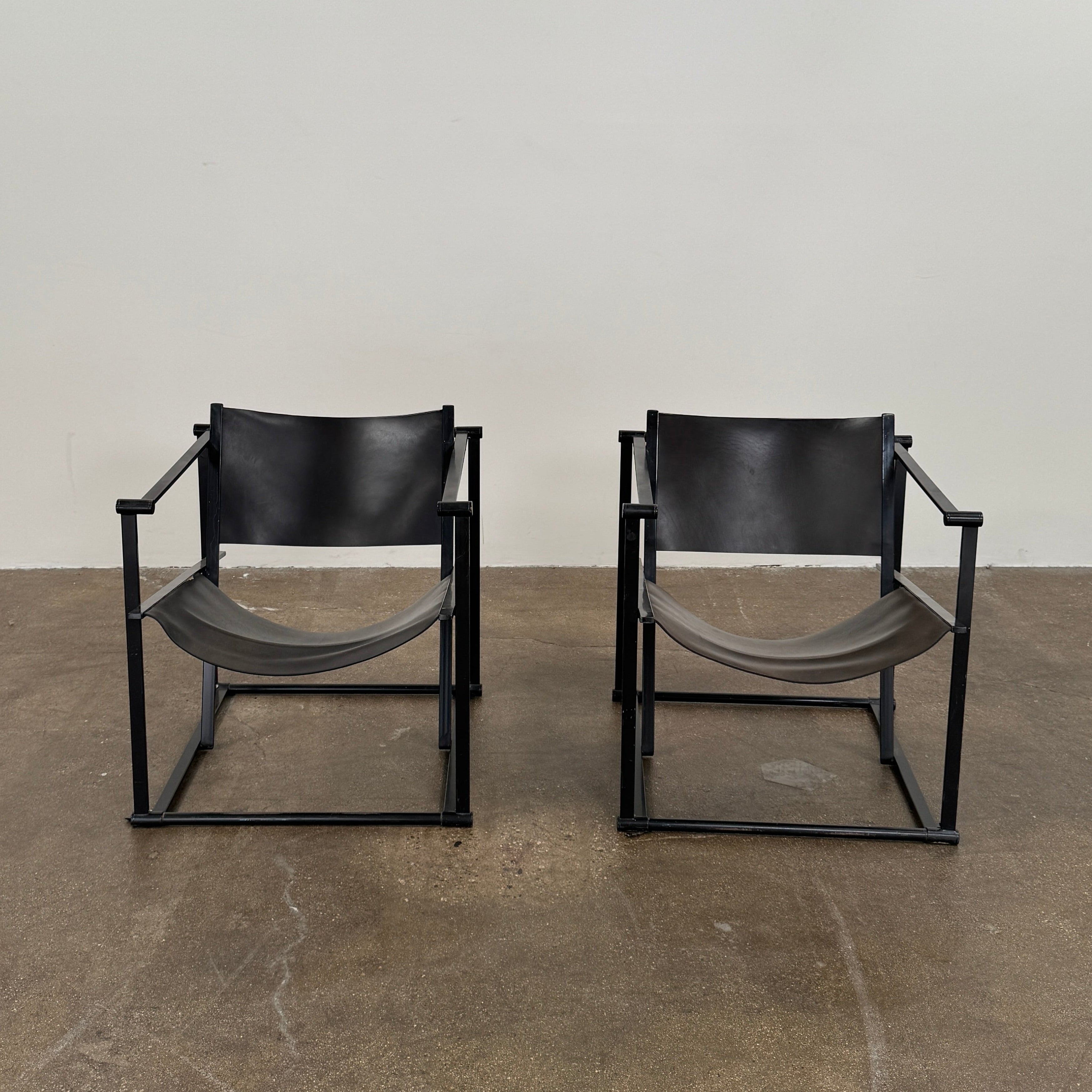 Two Pastoe FM62 chairs in black leather by Modern Resale are positioned side by side on a brown concrete floor against a plain white wall.