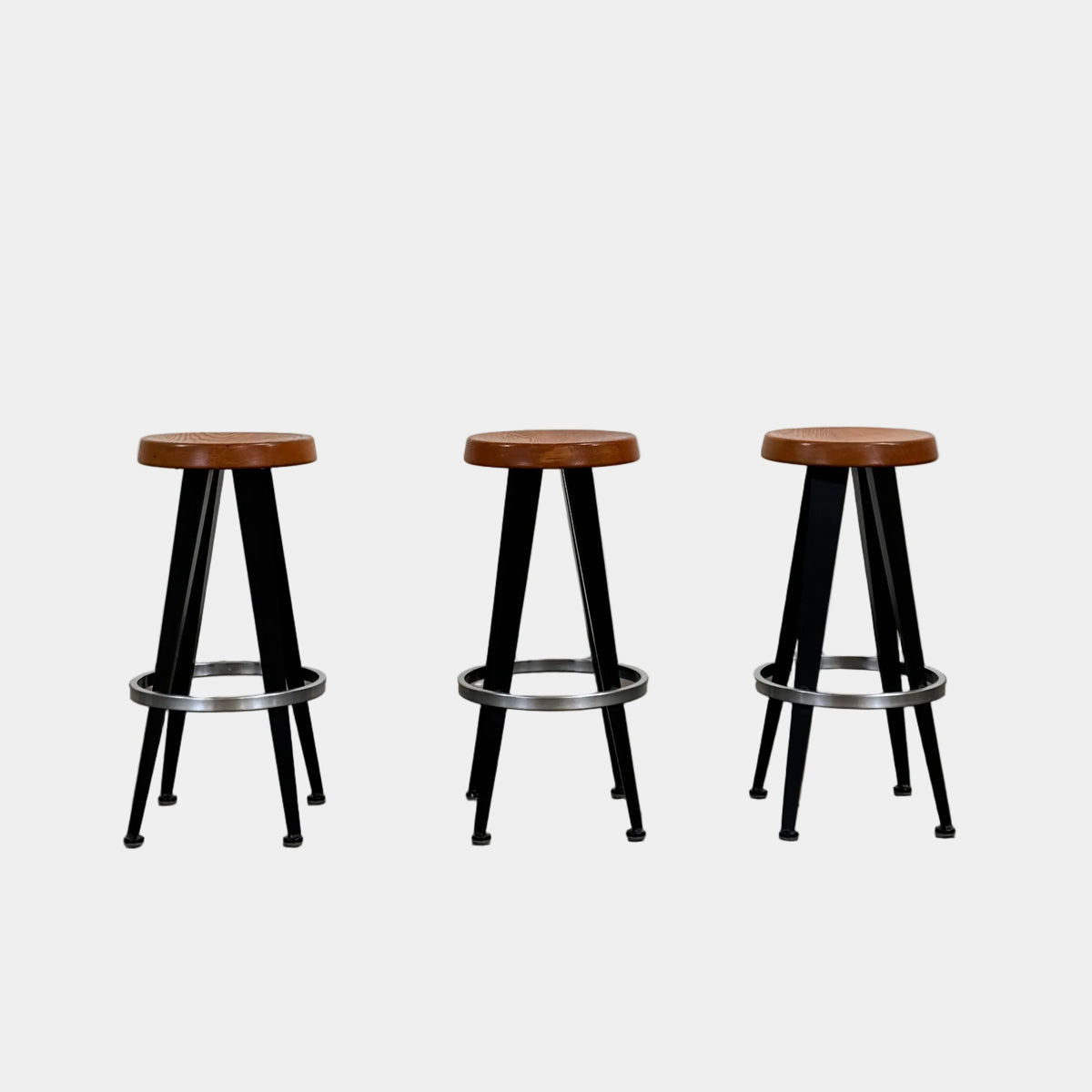 A set of three Vintage Stools in the style of Jean Prouve by Vintage, featuring black metal frames, round brown wooden seats, and silver footrests, are arranged in a row against a plain white background, evoking a classic mid-century design.