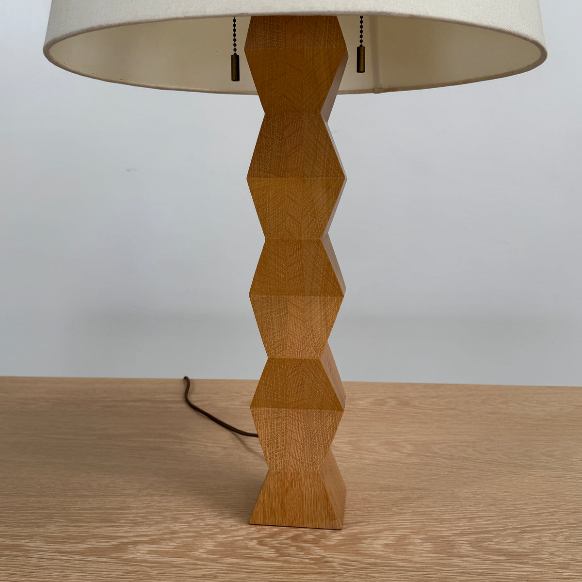 The Donghia Zig Zag Lamp Pickled Oak by Donghia features a white fabric shade and geometric wooden base, displayed on a light wood surface against a plain background.