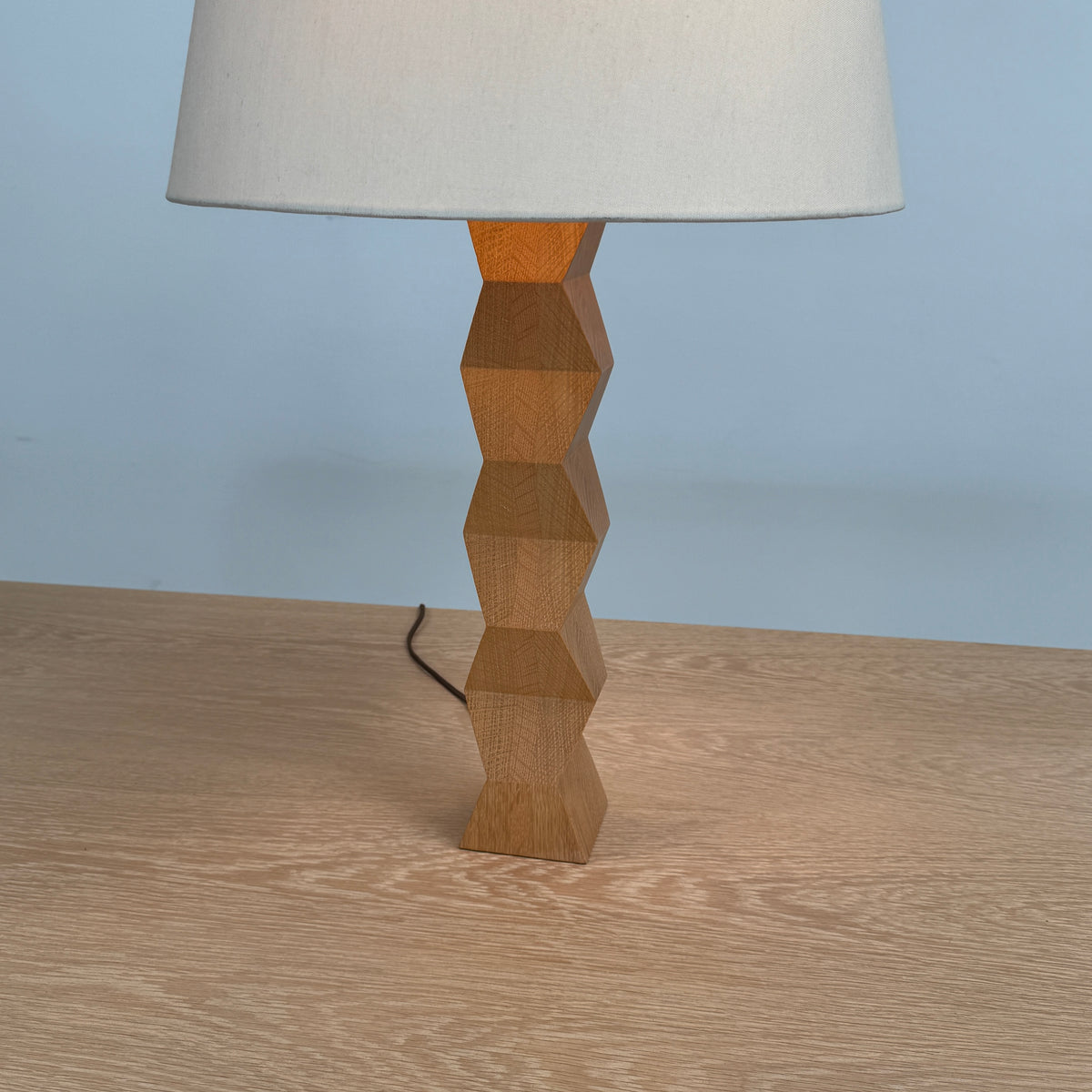 The Donghia Zig Zag Lamp Pickled Oak by Donghia features a white fabric shade and geometric wooden base, displayed on a light wood surface against a plain background.
