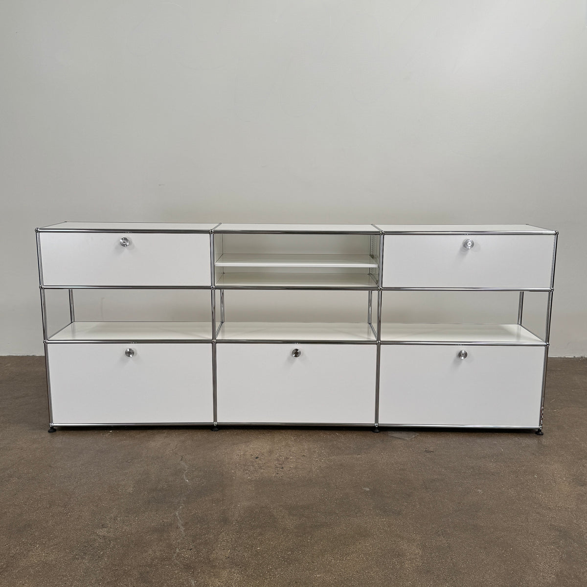 A USM Haller Storage Cabinet by USM, finished in minimal white, stands on a plain concrete floor against a light-colored wall. The modular cabinet features six closed compartments, two open shelves, and a sturdy metal frame.