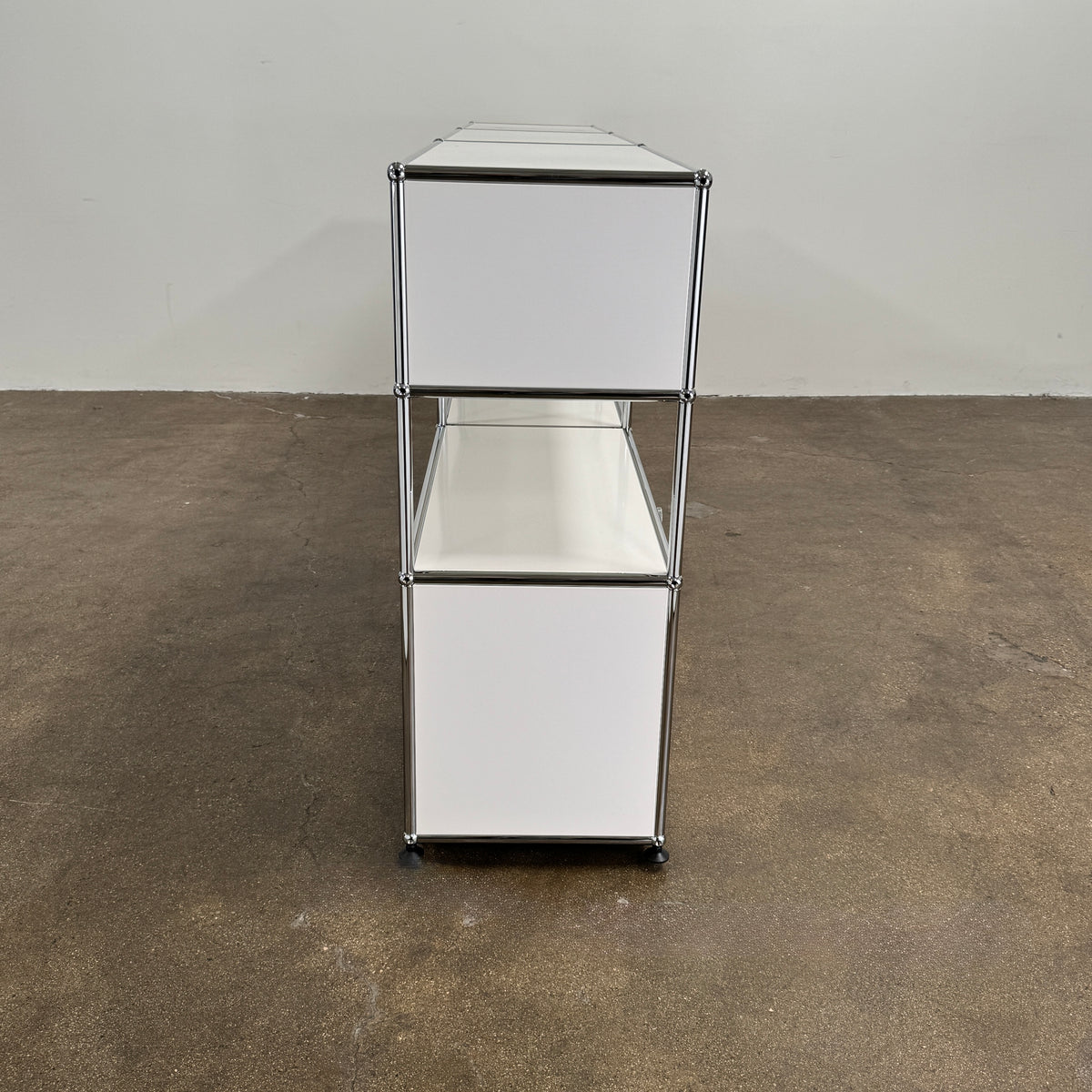 A USM Haller Storage Cabinet by USM, finished in minimal white, stands on a plain concrete floor against a light-colored wall. The modular cabinet features six closed compartments, two open shelves, and a sturdy metal frame.