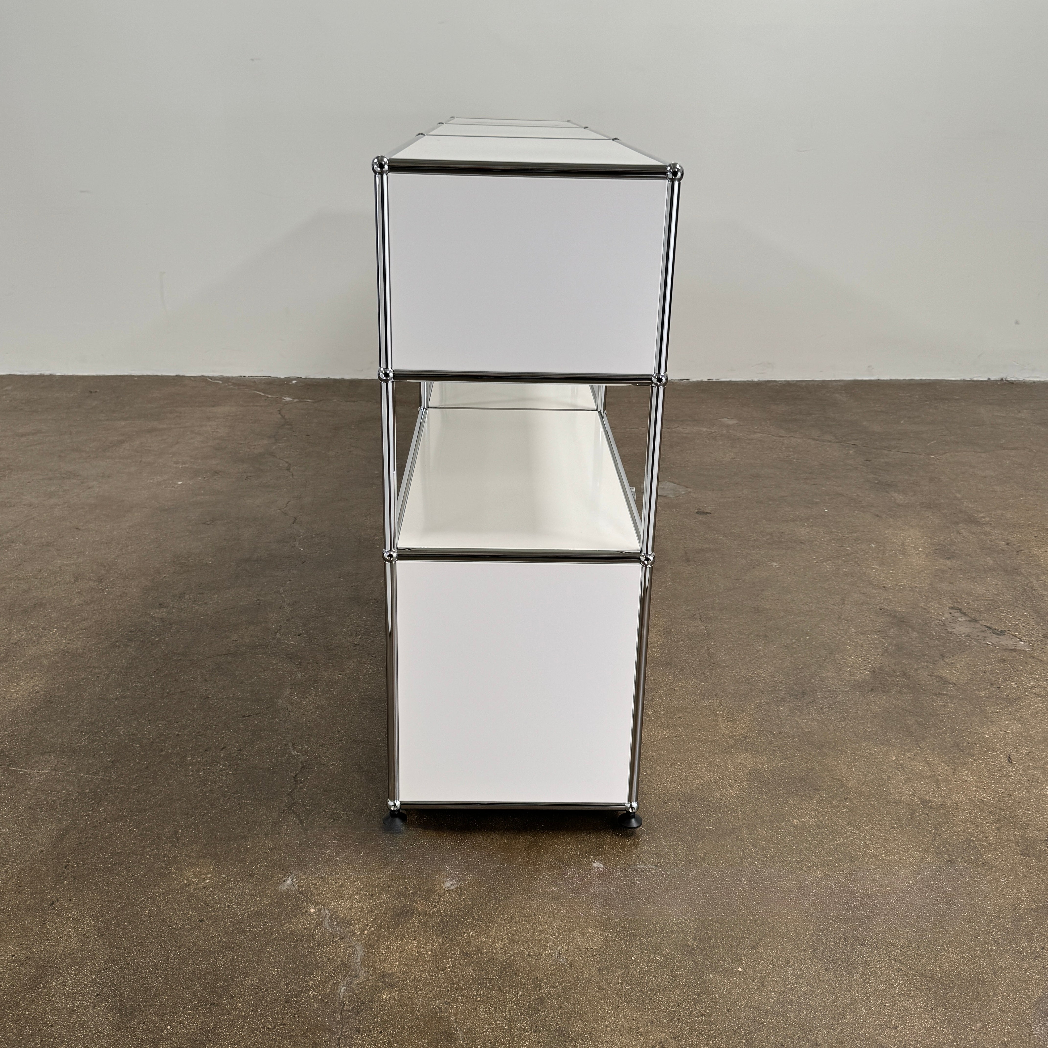 A USM Haller Storage Cabinet by USM, finished in minimal white, stands on a plain concrete floor against a light-colored wall. The modular cabinet features six closed compartments, two open shelves, and a sturdy metal frame.