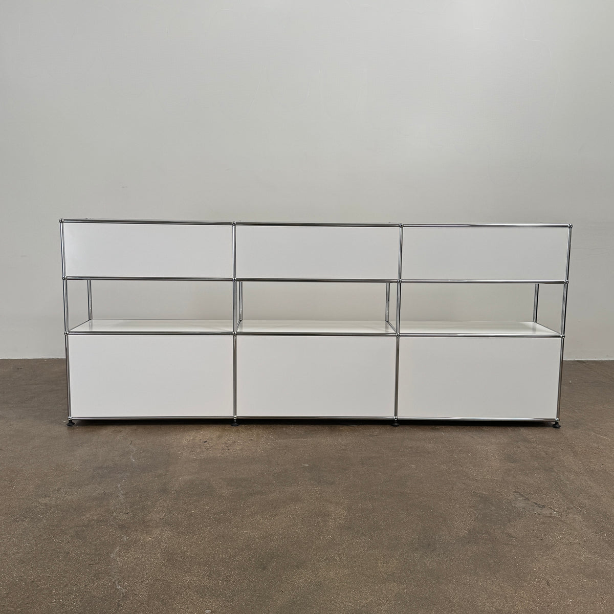 A USM Haller Storage Cabinet by USM, finished in minimal white, stands on a plain concrete floor against a light-colored wall. The modular cabinet features six closed compartments, two open shelves, and a sturdy metal frame.