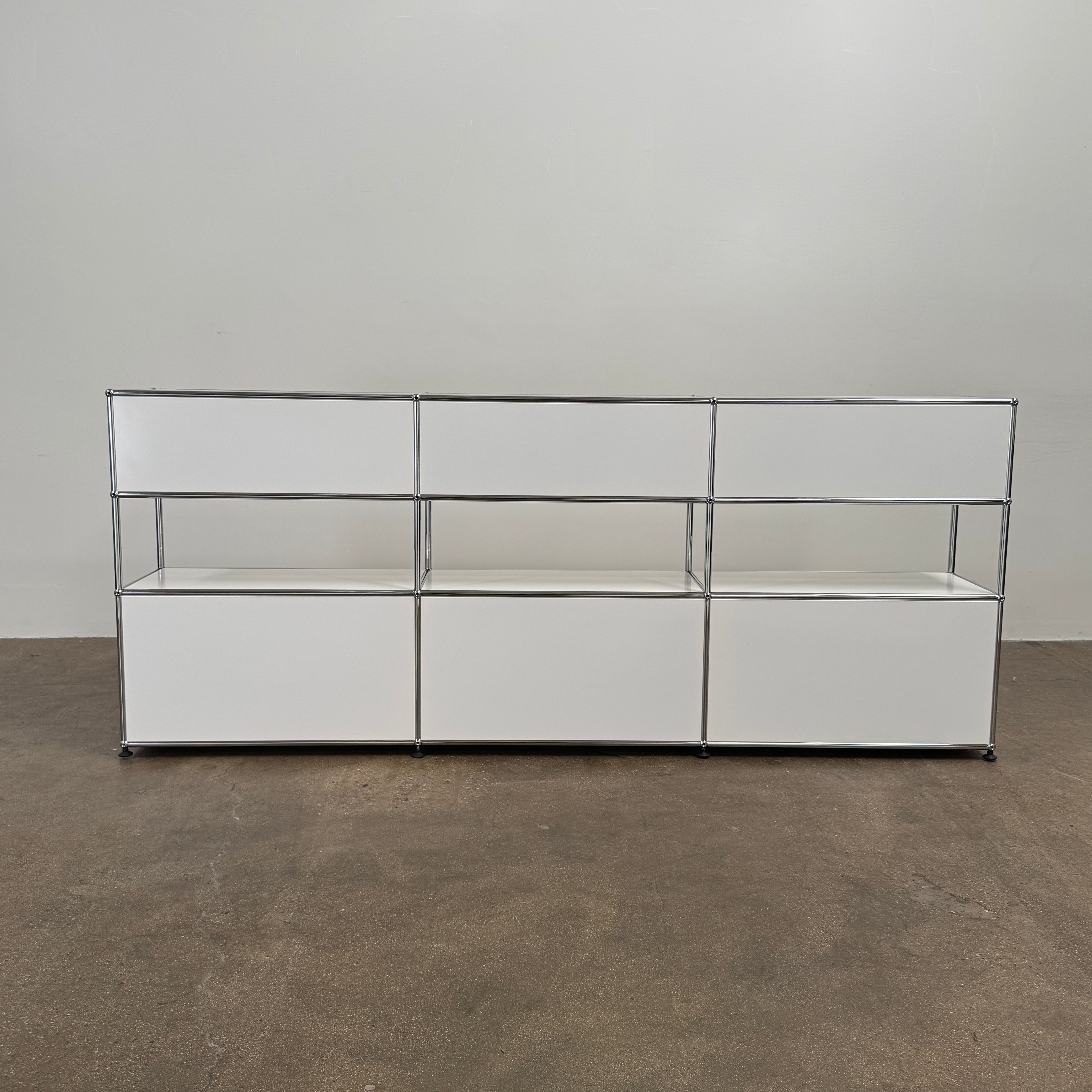 A USM Haller Storage Cabinet by USM, finished in minimal white, stands on a plain concrete floor against a light-colored wall. The modular cabinet features six closed compartments, two open shelves, and a sturdy metal frame.