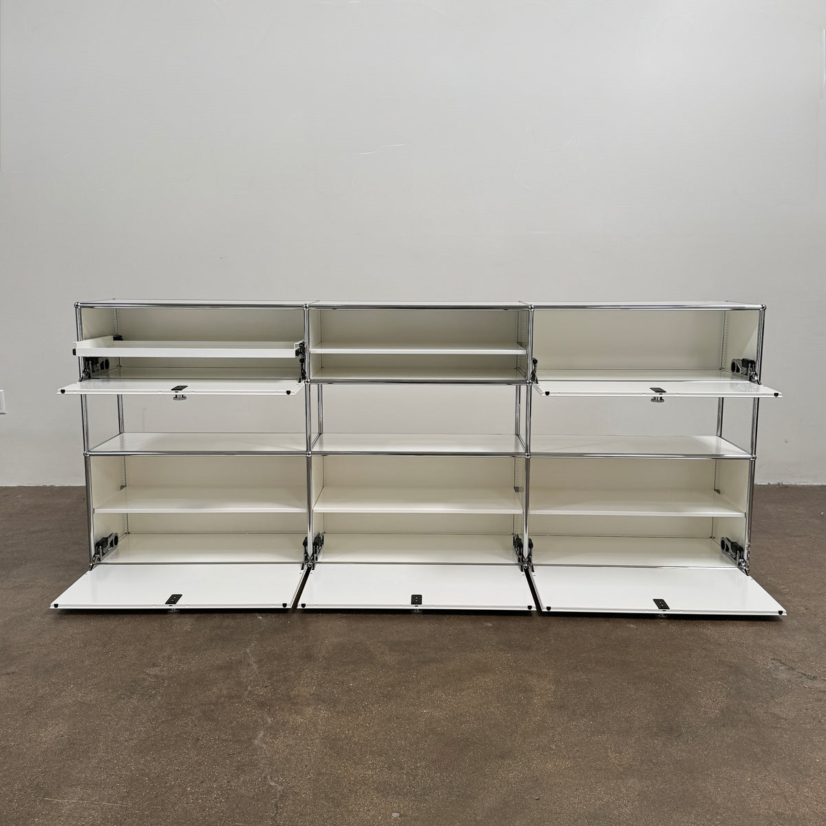 A USM Haller Storage Cabinet by USM, finished in minimal white, stands on a plain concrete floor against a light-colored wall. The modular cabinet features six closed compartments, two open shelves, and a sturdy metal frame.