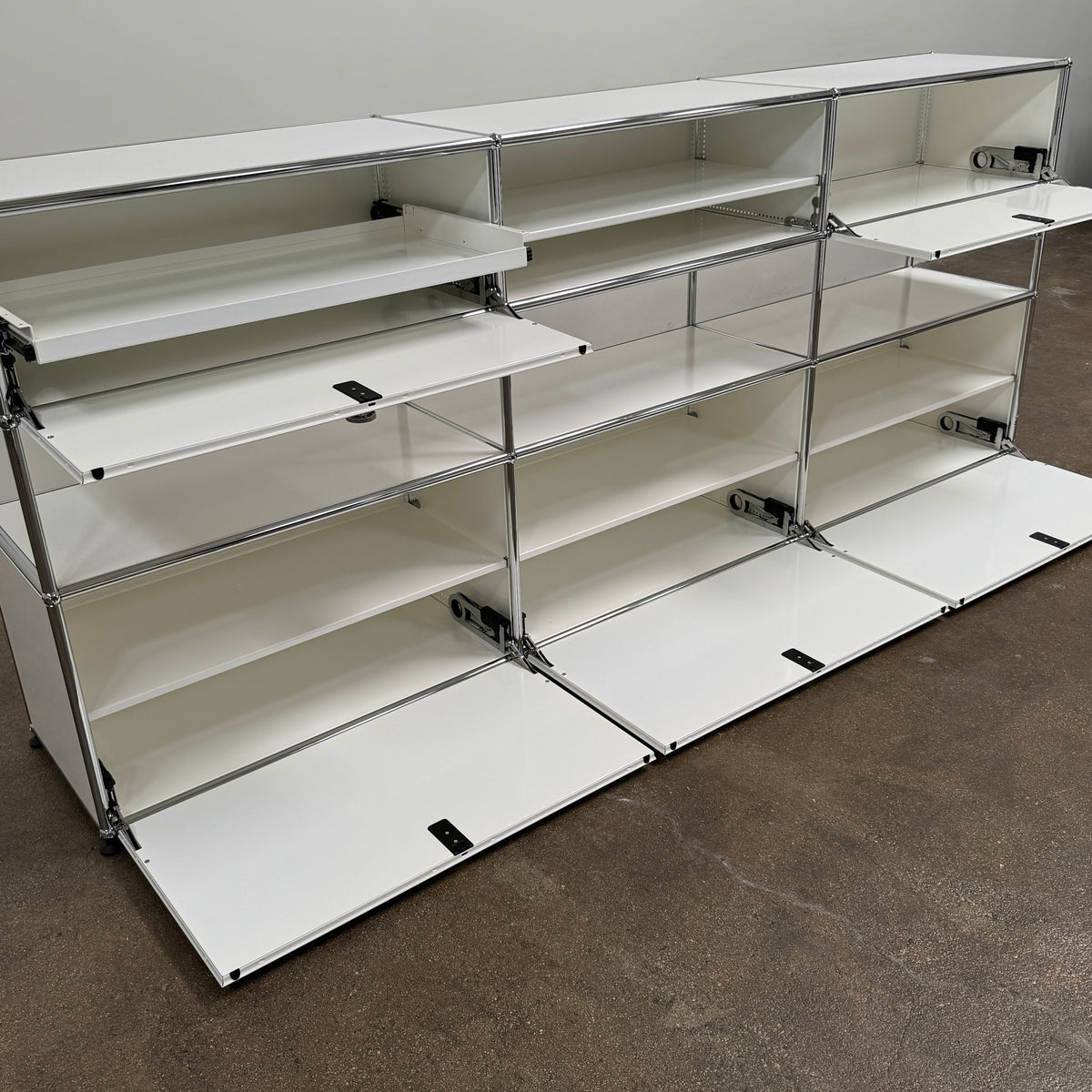 A USM Haller Storage Cabinet by USM, finished in minimal white, stands on a plain concrete floor against a light-colored wall. The modular cabinet features six closed compartments, two open shelves, and a sturdy metal frame.