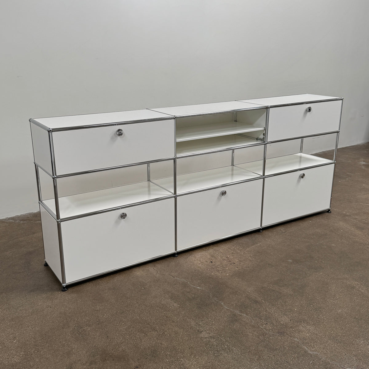 A USM Haller Storage Cabinet by USM, finished in minimal white, stands on a plain concrete floor against a light-colored wall. The modular cabinet features six closed compartments, two open shelves, and a sturdy metal frame.