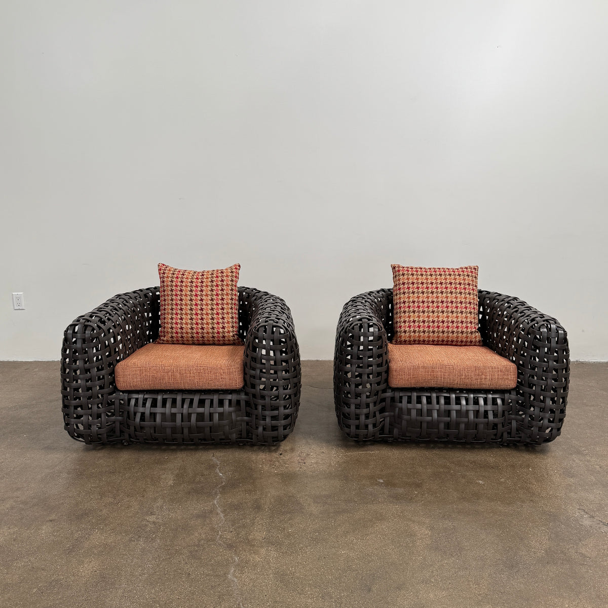 Two Outdoor Matilda Easy Armchairs by Kenneth Cobonpue from Modern Resale, featuring dark woven frames with orange patterned seat and back cushions, sit side by side on a brown concrete floor against a plain white wall.