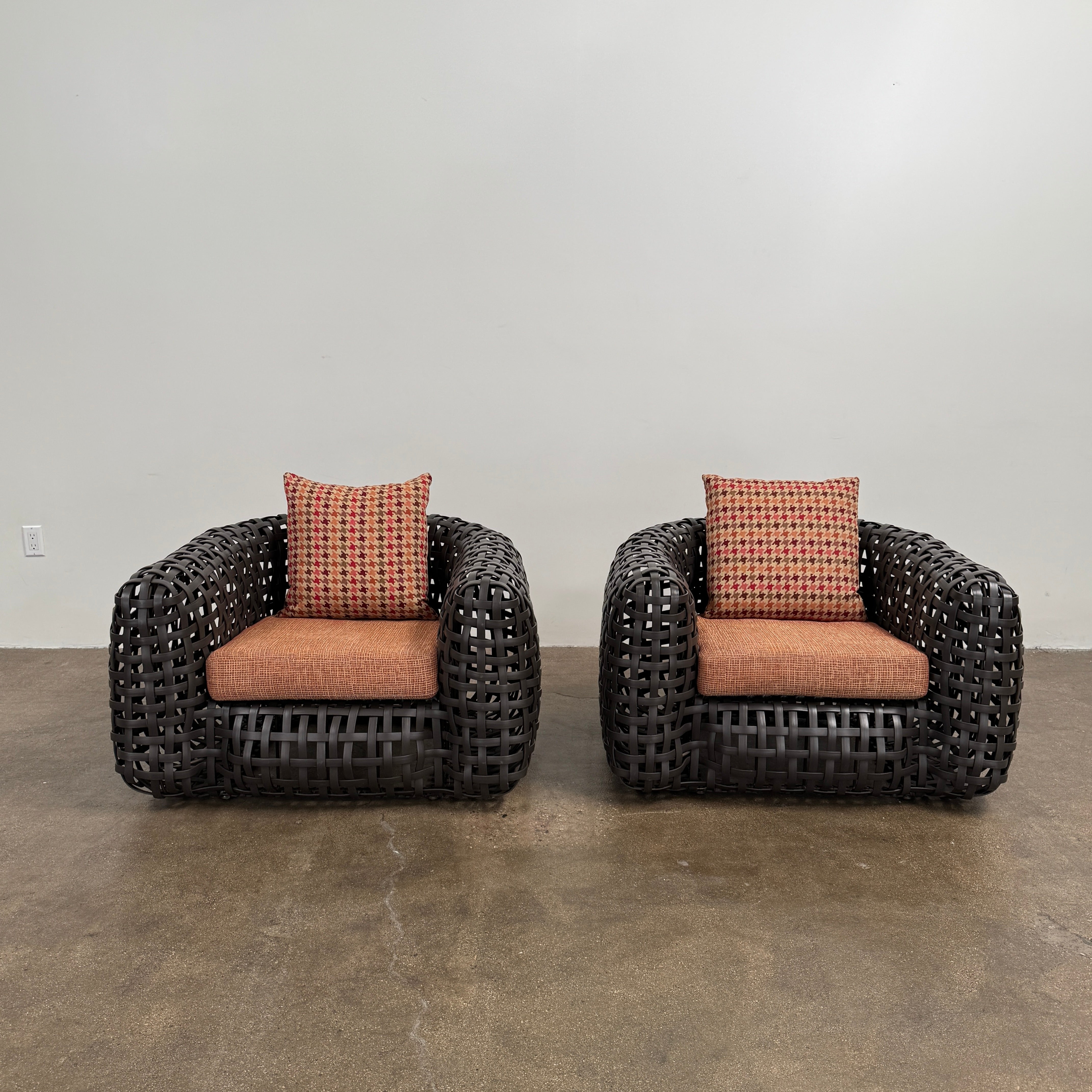 Two Outdoor Matilda Easy Armchairs by Kenneth Cobonpue from Modern Resale, featuring dark woven frames with orange patterned seat and back cushions, sit side by side on a brown concrete floor against a plain white wall.