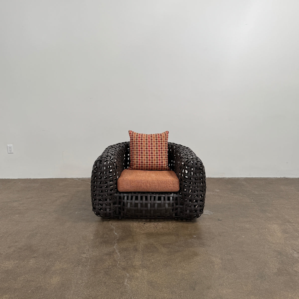 Two Outdoor Matilda Easy Armchairs by Kenneth Cobonpue from Modern Resale, featuring dark woven frames with orange patterned seat and back cushions, sit side by side on a brown concrete floor against a plain white wall.