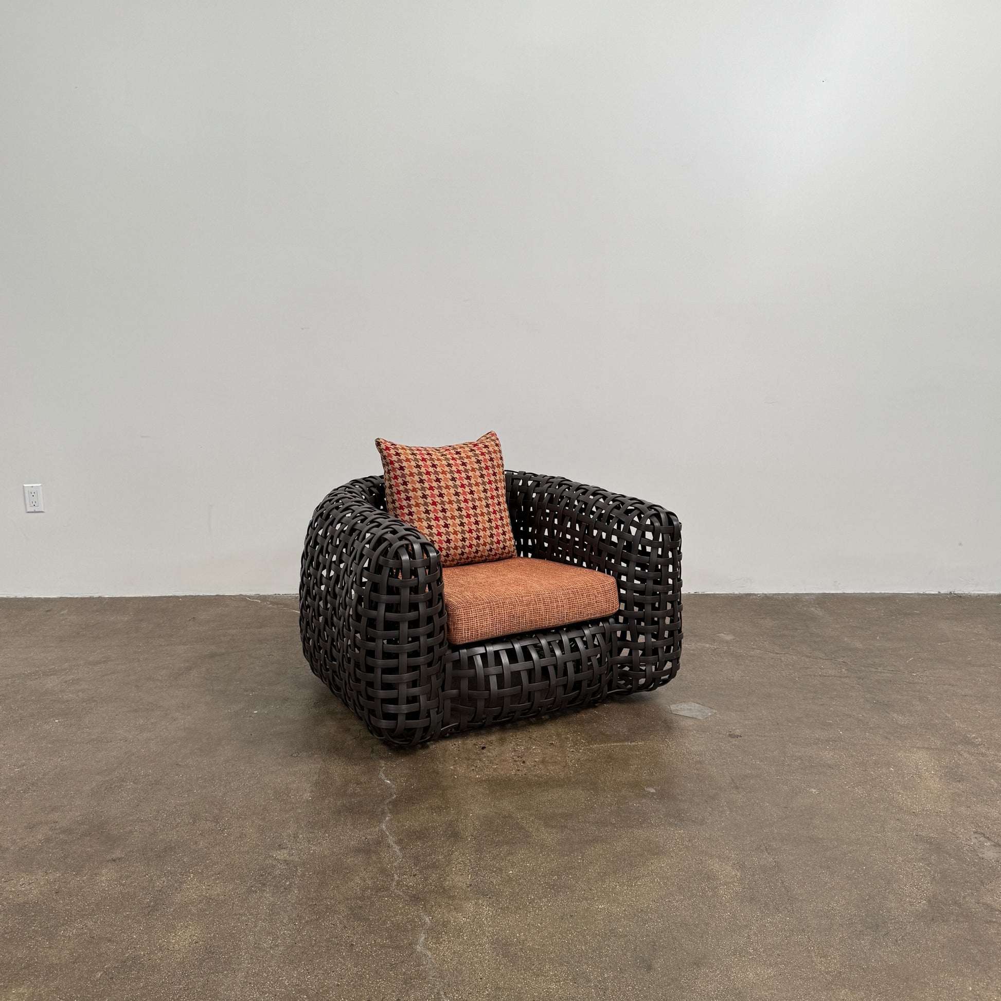 Two Outdoor Matilda Easy Armchairs by Kenneth Cobonpue from Modern Resale, featuring dark woven frames with orange patterned seat and back cushions, sit side by side on a brown concrete floor against a plain white wall.