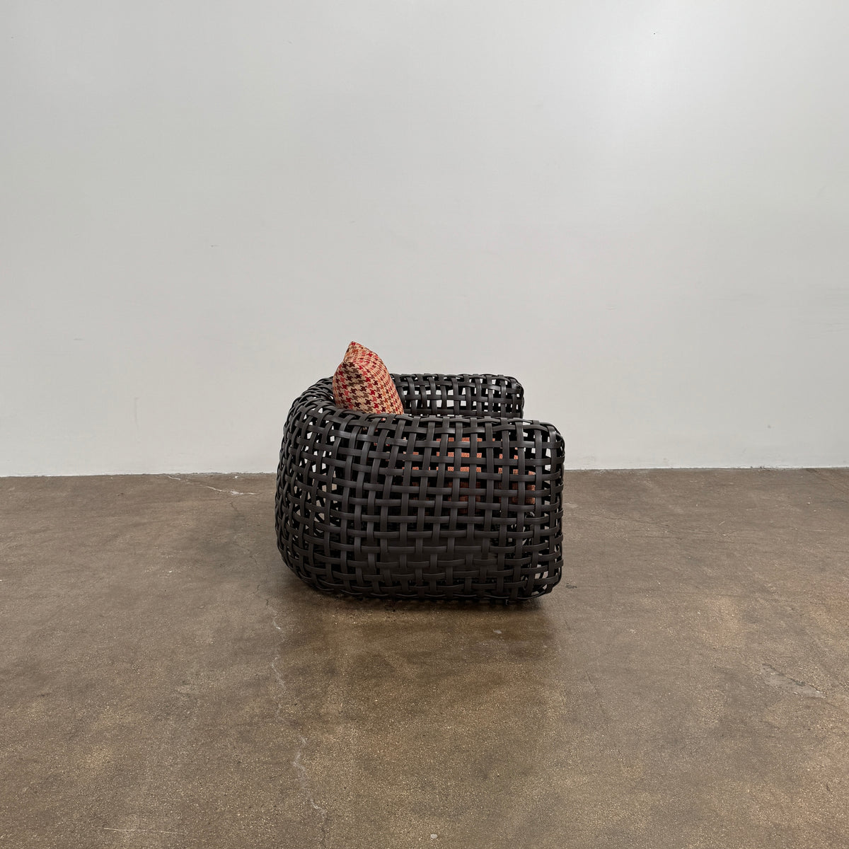 Two Outdoor Matilda Easy Armchairs by Kenneth Cobonpue from Modern Resale, featuring dark woven frames with orange patterned seat and back cushions, sit side by side on a brown concrete floor against a plain white wall.