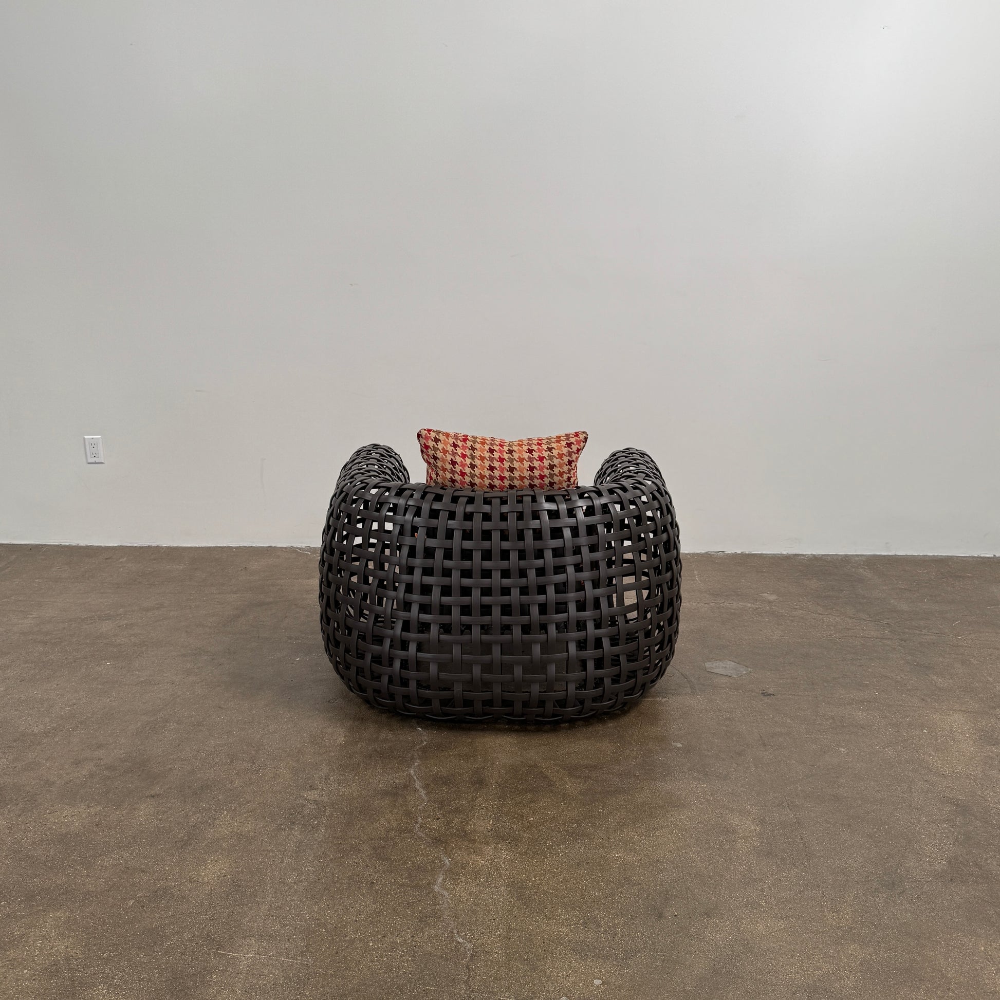 Two Outdoor Matilda Easy Armchairs by Kenneth Cobonpue from Modern Resale, featuring dark woven frames with orange patterned seat and back cushions, sit side by side on a brown concrete floor against a plain white wall.