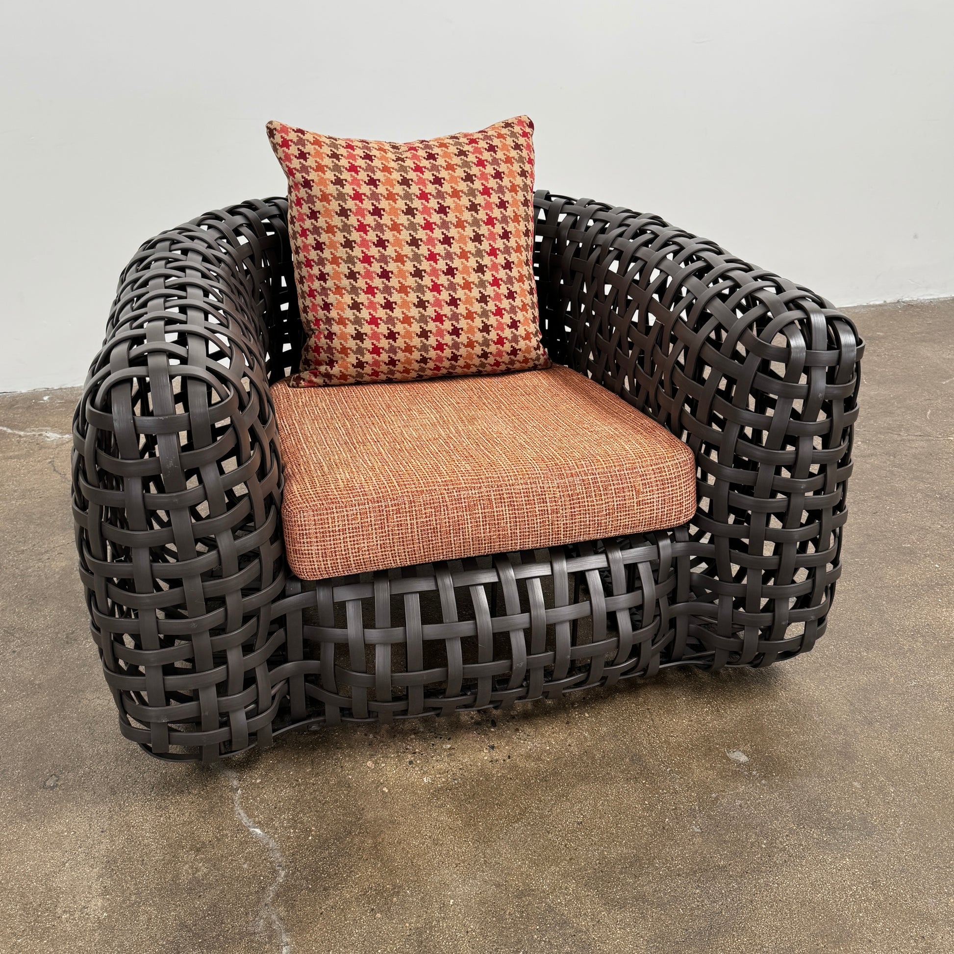 Two Outdoor Matilda Easy Armchairs by Kenneth Cobonpue from Modern Resale, featuring dark woven frames with orange patterned seat and back cushions, sit side by side on a brown concrete floor against a plain white wall.