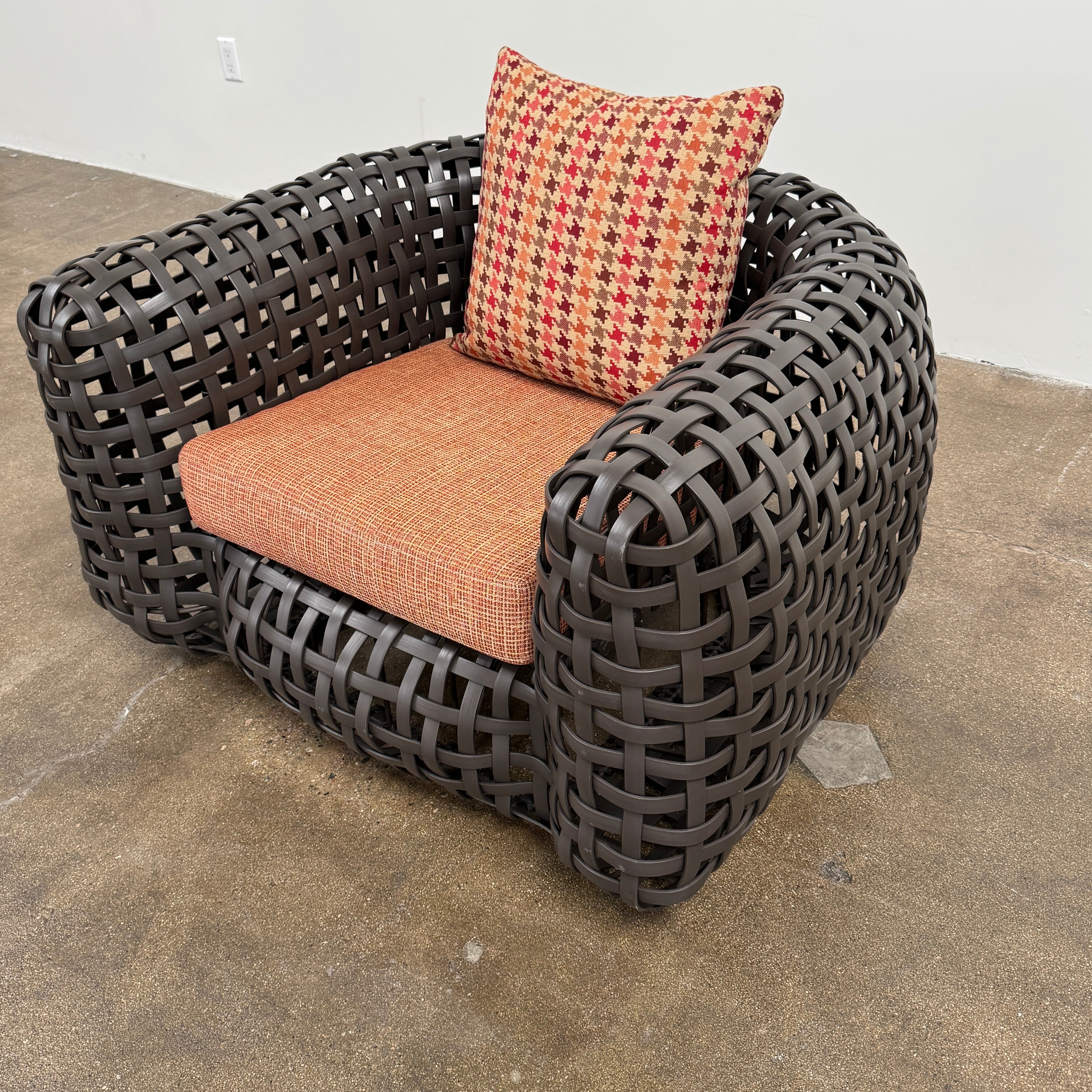 Two Outdoor Matilda Easy Armchairs by Kenneth Cobonpue from Modern Resale, featuring dark woven frames with orange patterned seat and back cushions, sit side by side on a brown concrete floor against a plain white wall.