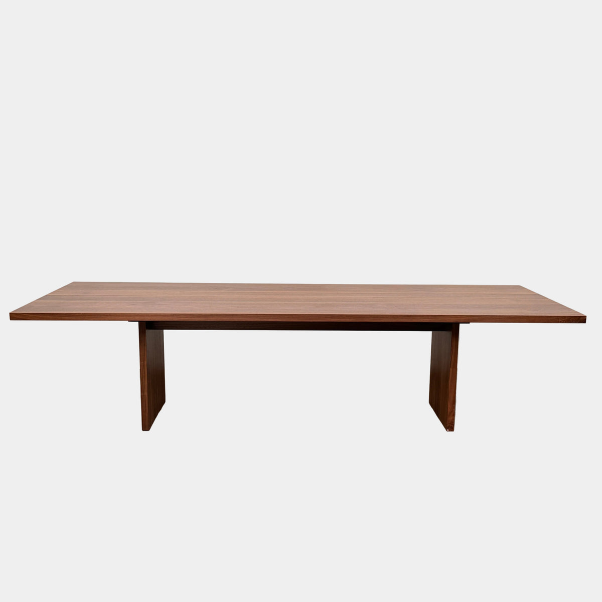 The DK3 Gather Table - Walnut by Dk3 features a rectangular shape with a split plank design, elevated on a single central pedestal base and crafted from solid walnut; it is displayed on a polished concrete floor against a plain white wall.