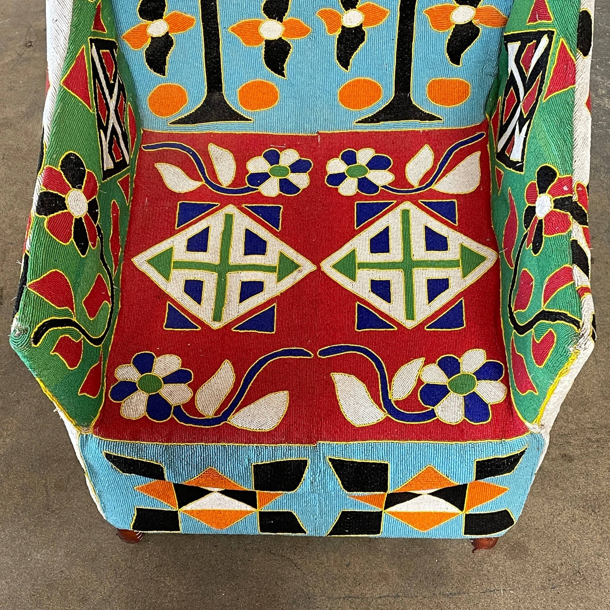 The Handmade Hand Beaded African Yoruba Chair (on hold) features a high backrest, wooden legs, and vibrant upholstery with multicolored geometric and floral patterns inspired by West African artistry.