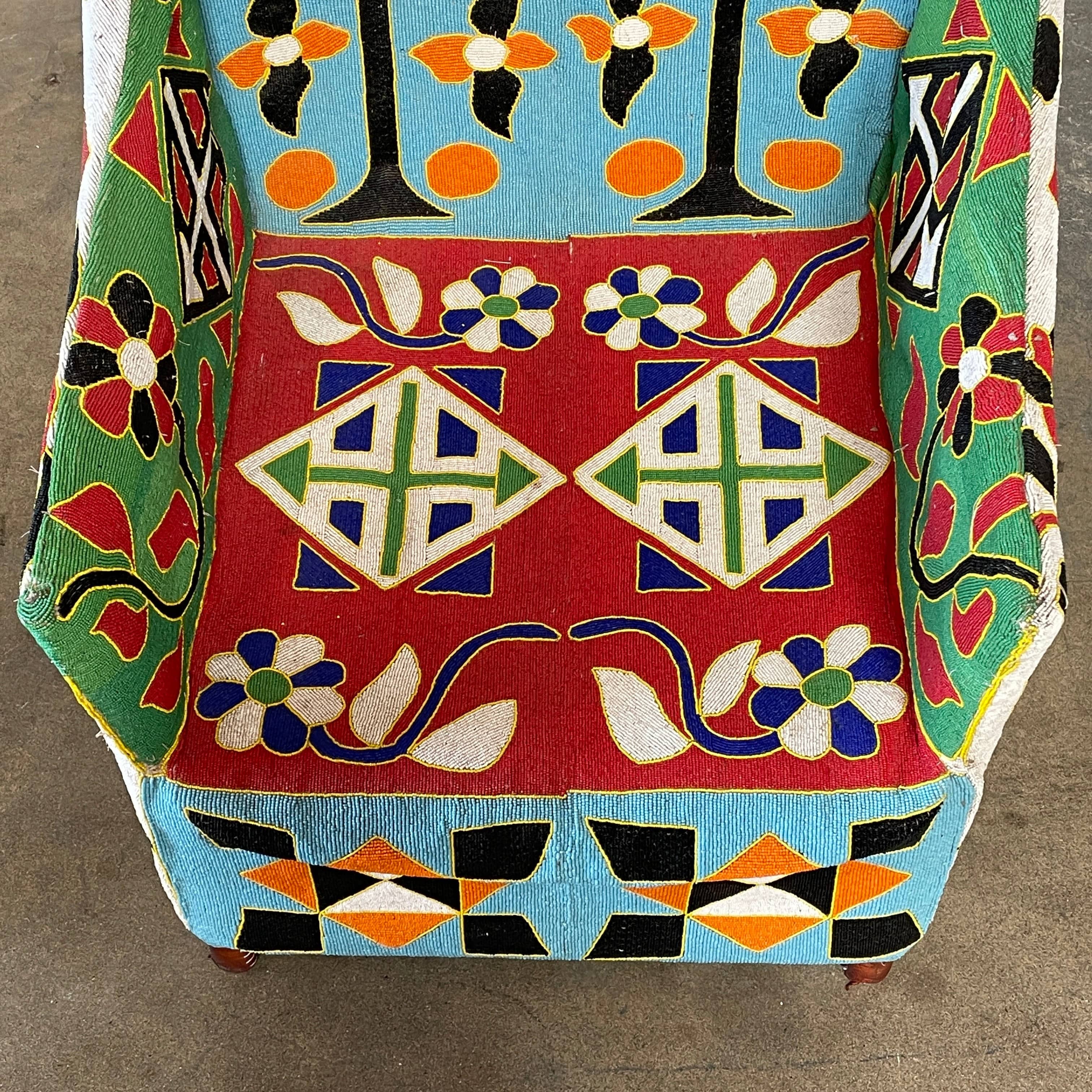 The Handmade Hand Beaded African Yoruba Chair (on hold) features a high backrest, wooden legs, and vibrant upholstery with multicolored geometric and floral patterns inspired by West African artistry.