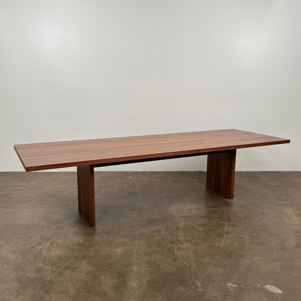 The DK3 Gather Table - Walnut by Dk3 features a rectangular shape with a split plank design, elevated on a single central pedestal base and crafted from solid walnut; it is displayed on a polished concrete floor against a plain white wall.