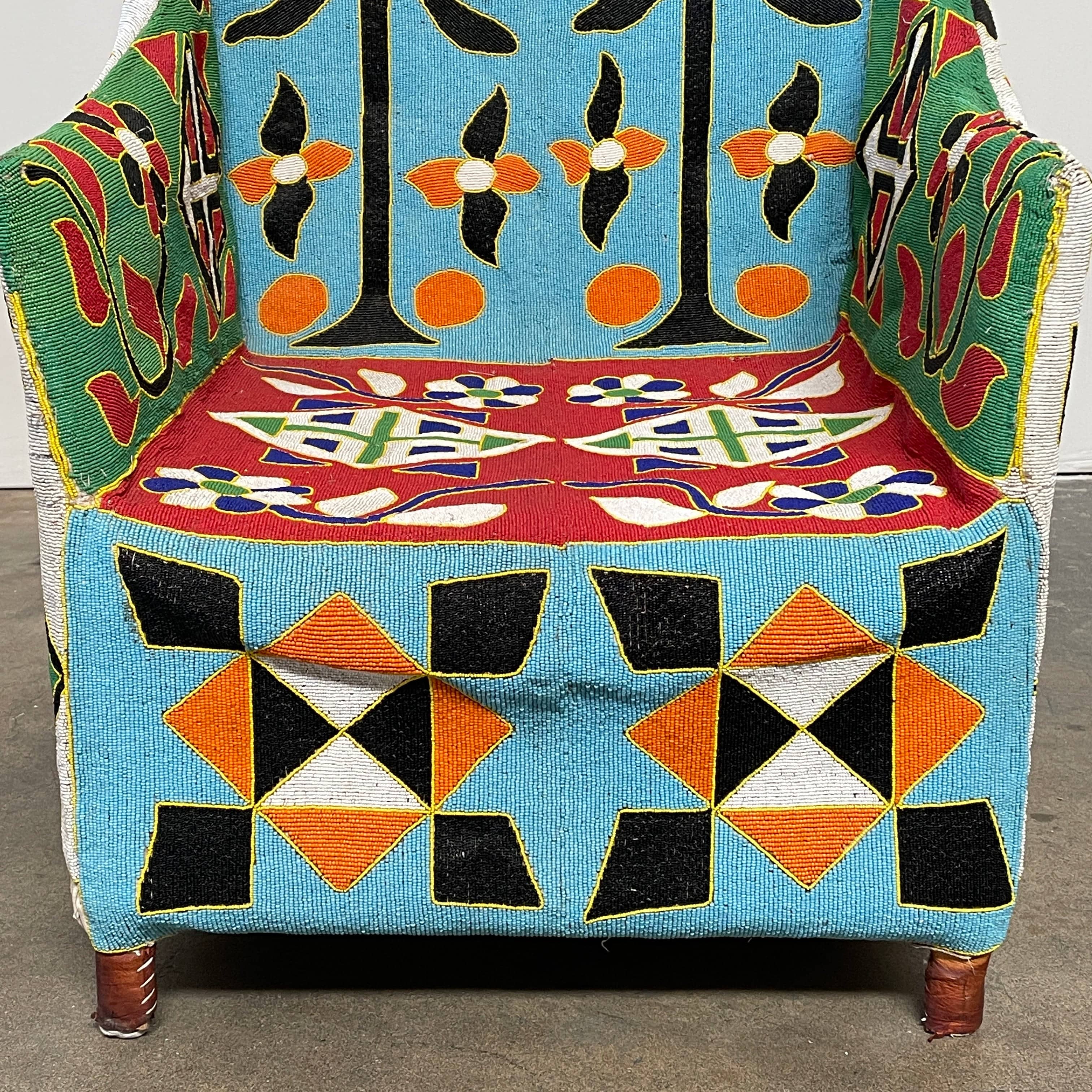 The Handmade Hand Beaded African Yoruba Chair (on hold) features a high backrest, wooden legs, and vibrant upholstery with multicolored geometric and floral patterns inspired by West African artistry.