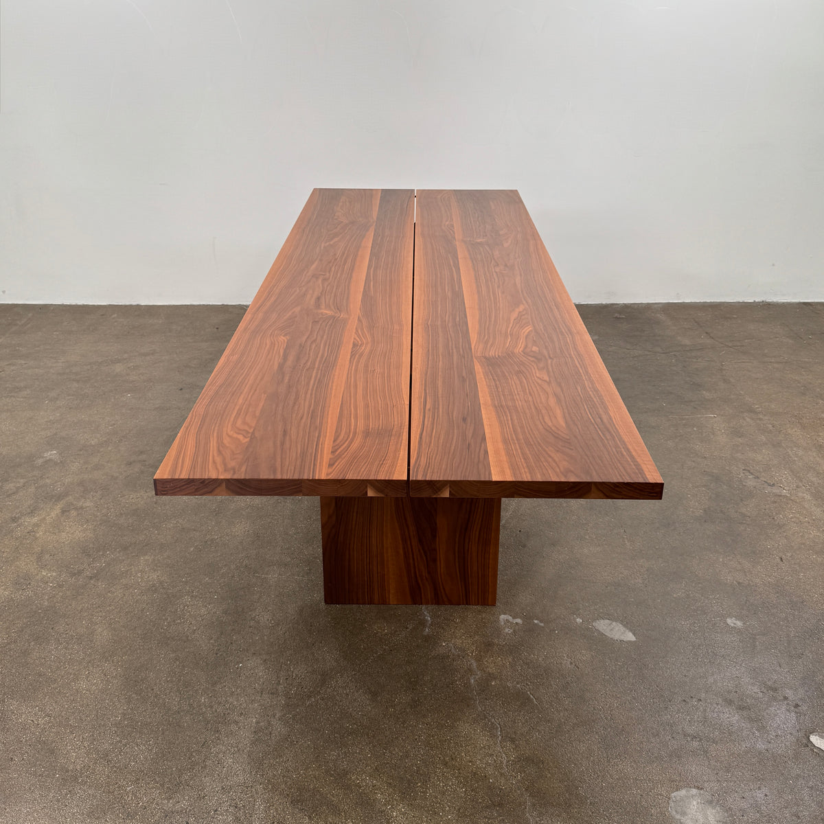 The DK3 Gather Table - Walnut by Dk3 features a rectangular shape with a split plank design, elevated on a single central pedestal base and crafted from solid walnut; it is displayed on a polished concrete floor against a plain white wall.