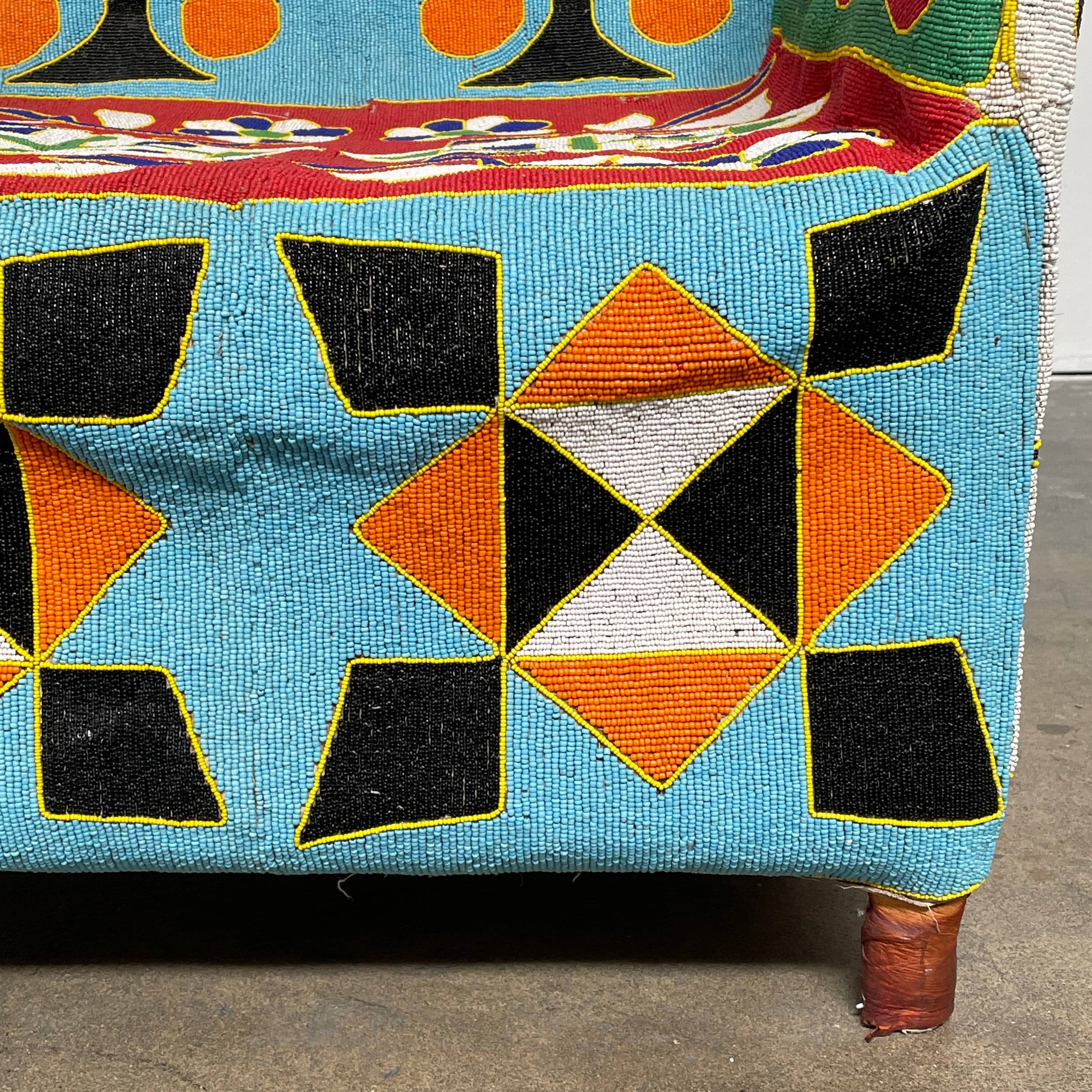 The Handmade Hand Beaded African Yoruba Chair (on hold) features a high backrest, wooden legs, and vibrant upholstery with multicolored geometric and floral patterns inspired by West African artistry.