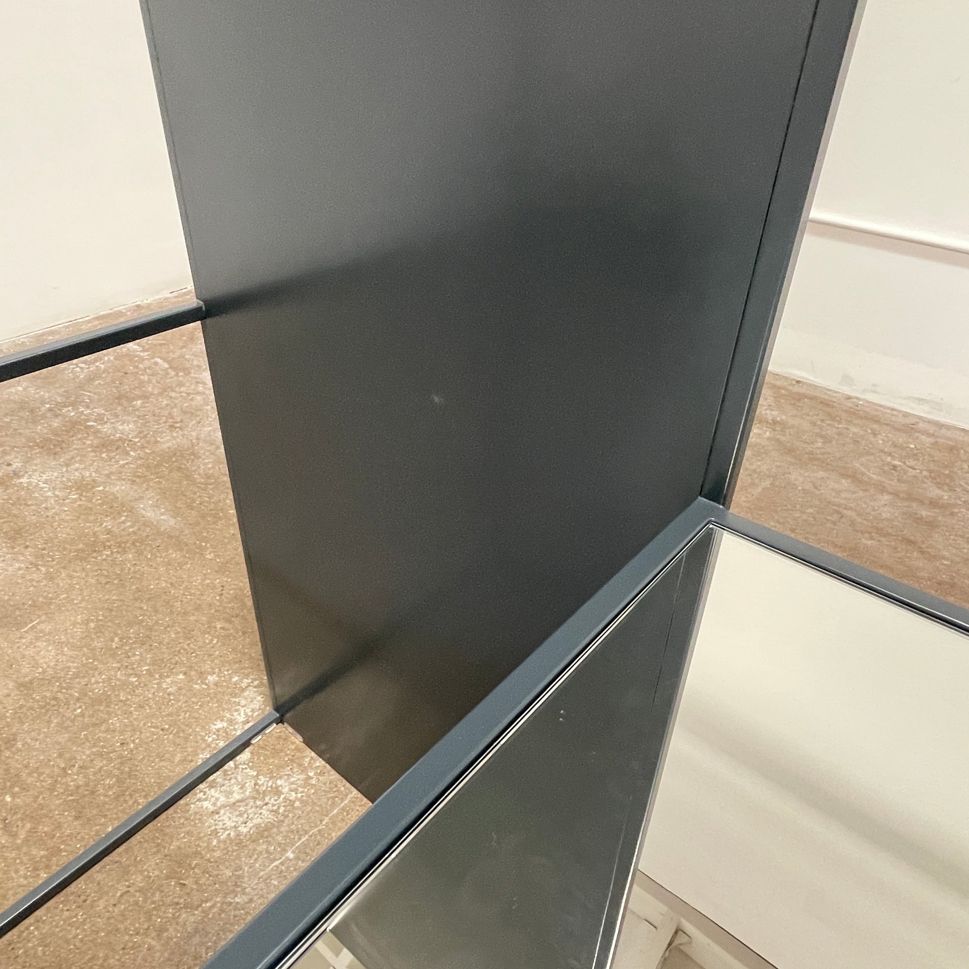 Tall, rectangular Cappellini Waku Mirrored Clothing Rack with a black metal frame, reflecting a room with a gray sofa, a colorful painting, and a beige carpet.