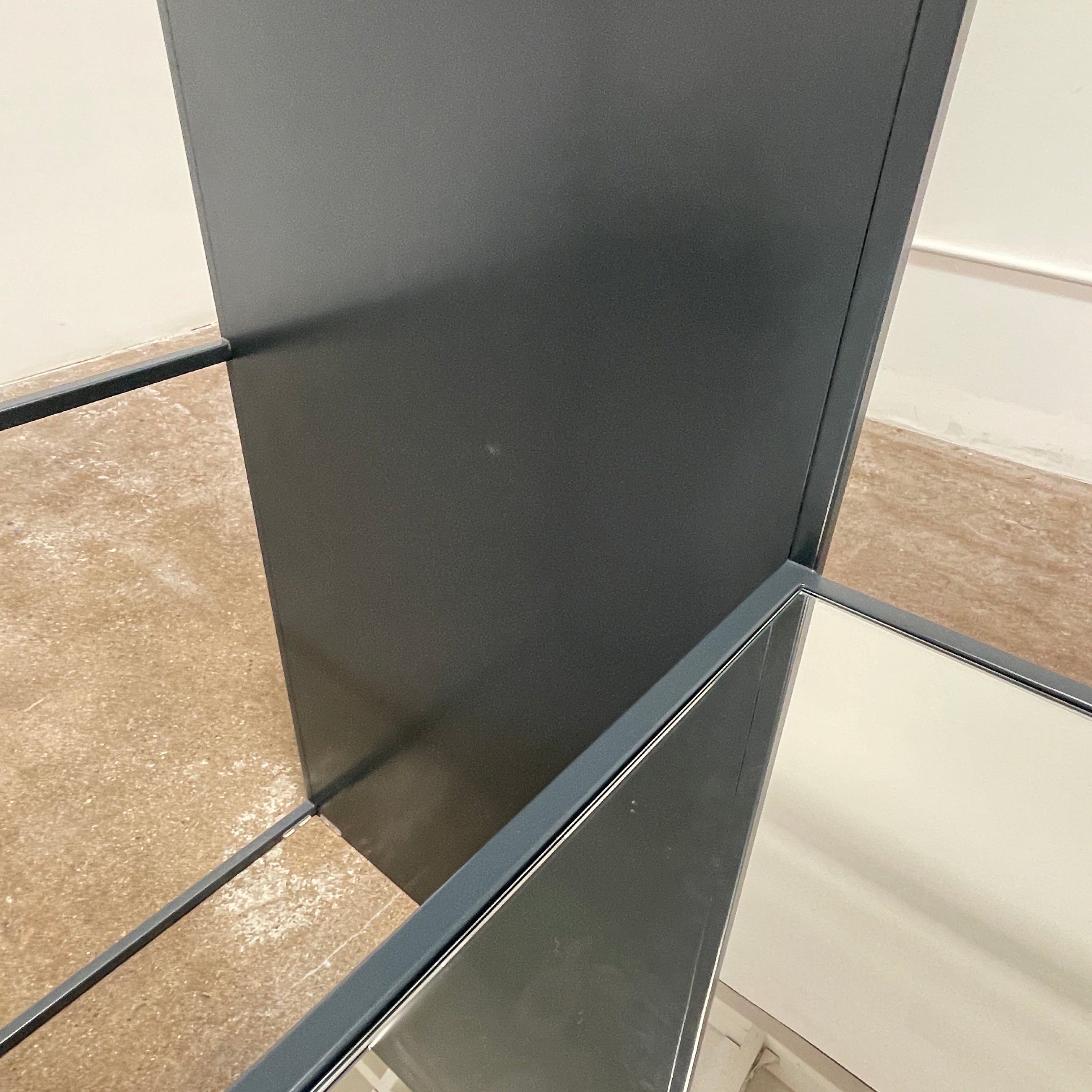 Tall, rectangular Cappellini Waku Mirrored Clothing Rack with a black metal frame, reflecting a room with a gray sofa, a colorful painting, and a beige carpet.