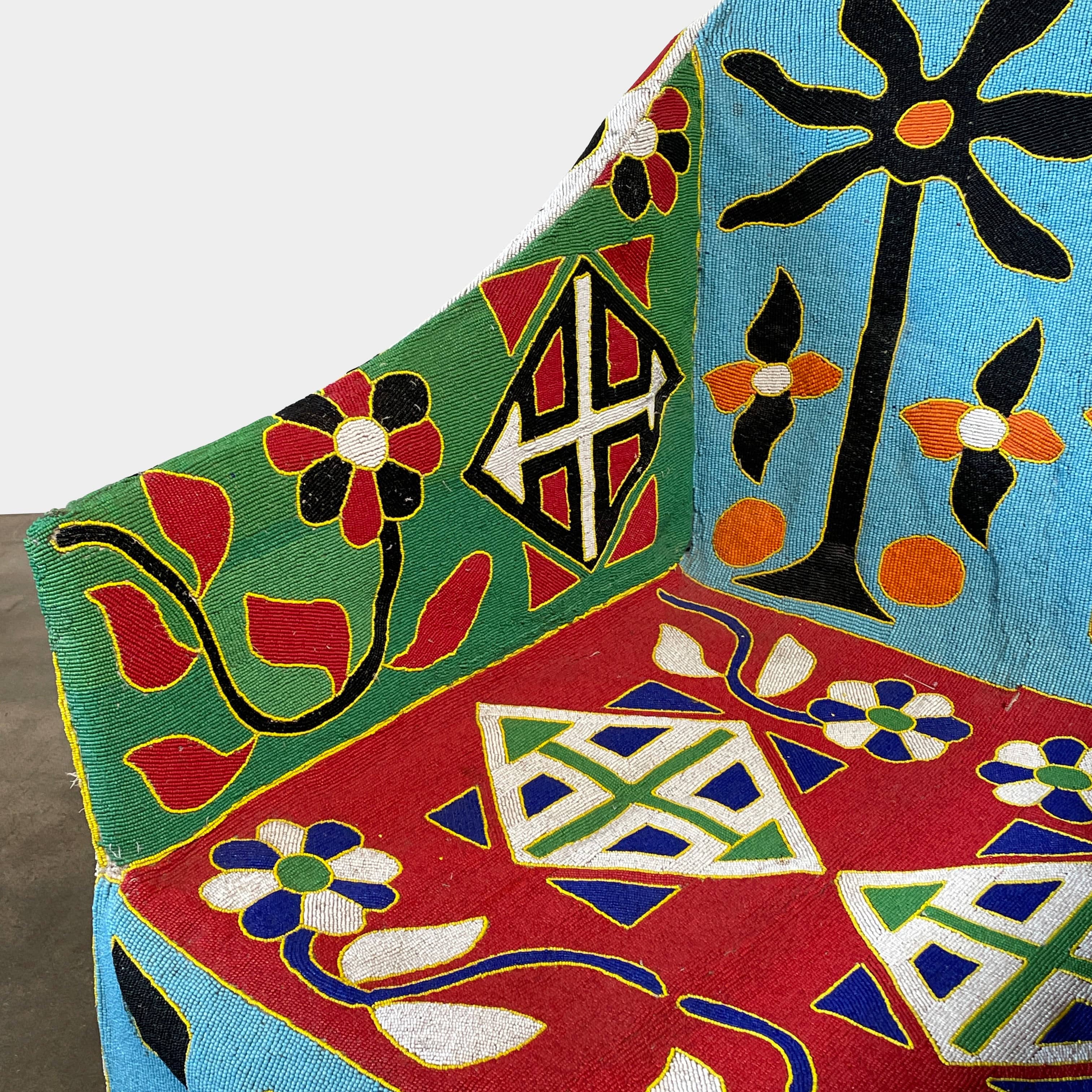 The Handmade Hand Beaded African Yoruba Chair (on hold) features a high backrest, wooden legs, and vibrant upholstery with multicolored geometric and floral patterns inspired by West African artistry.