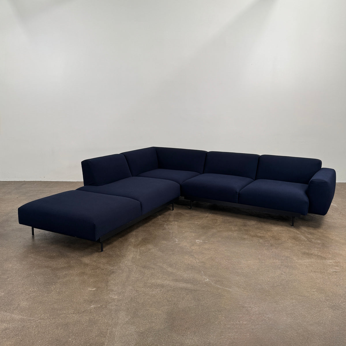 The Muuto Outline 3 Piece Sectional Sofa by Muuto features a dark blue fabric upholstery in a sleek, L-shaped design with low backrests and black legs, echoing the clean lines of contemporary architecture. The sectional stands out beautifully against a plain white background.