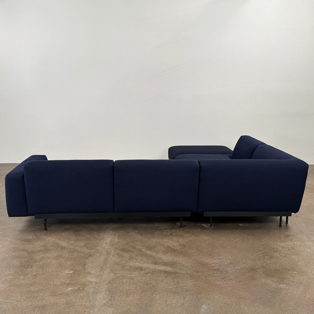 The Muuto Outline 3 Piece Sectional Sofa by Muuto features a dark blue fabric upholstery in a sleek, L-shaped design with low backrests and black legs, echoing the clean lines of contemporary architecture. The sectional stands out beautifully against a plain white background.