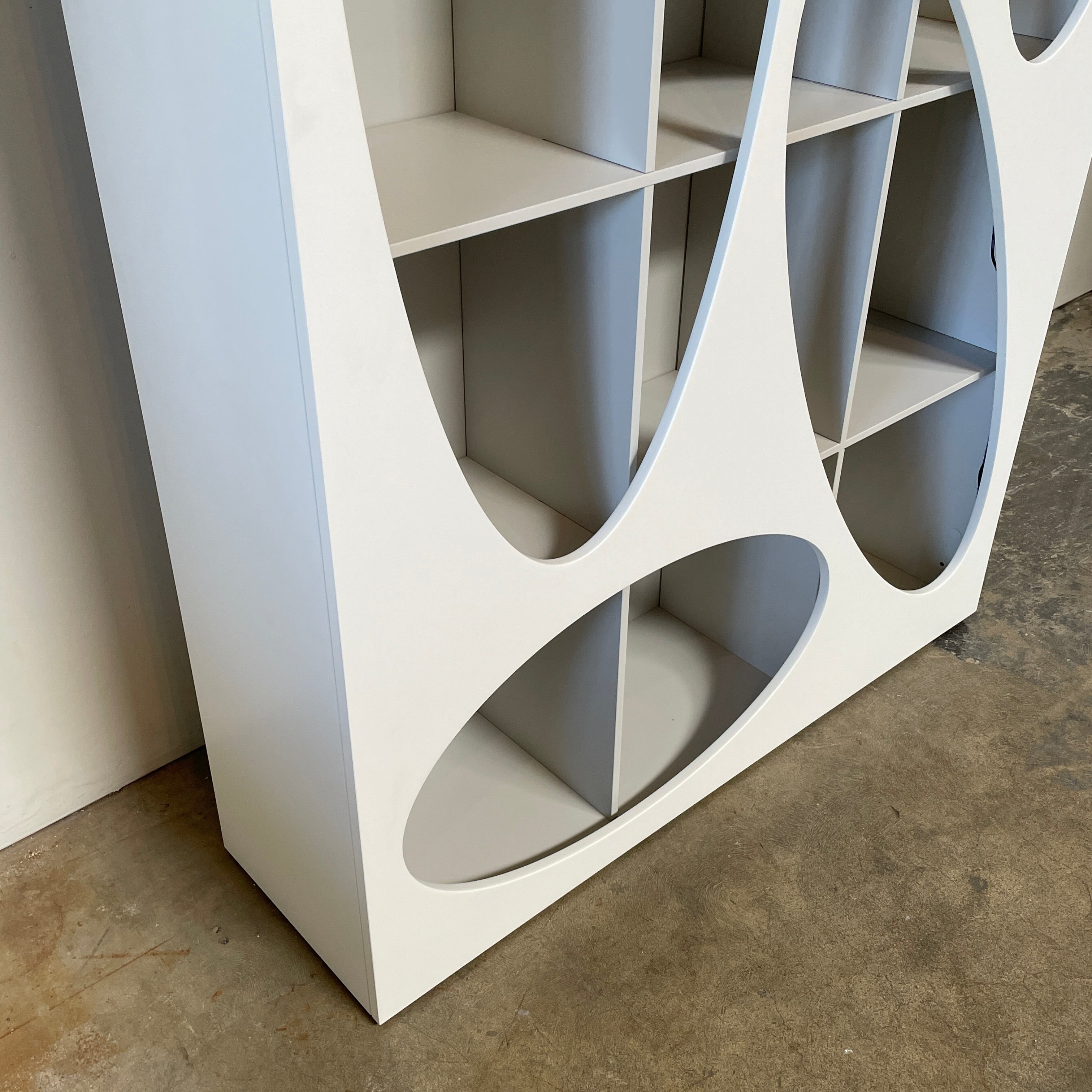 The Roche Bobois Denia Bookcase is a white bookshelf characterized by its abstract design, featuring multiple irregular oval cutouts, nine internal compartments, and integrated LED lighting, reminiscent of ROCHE BOBOIS styling.