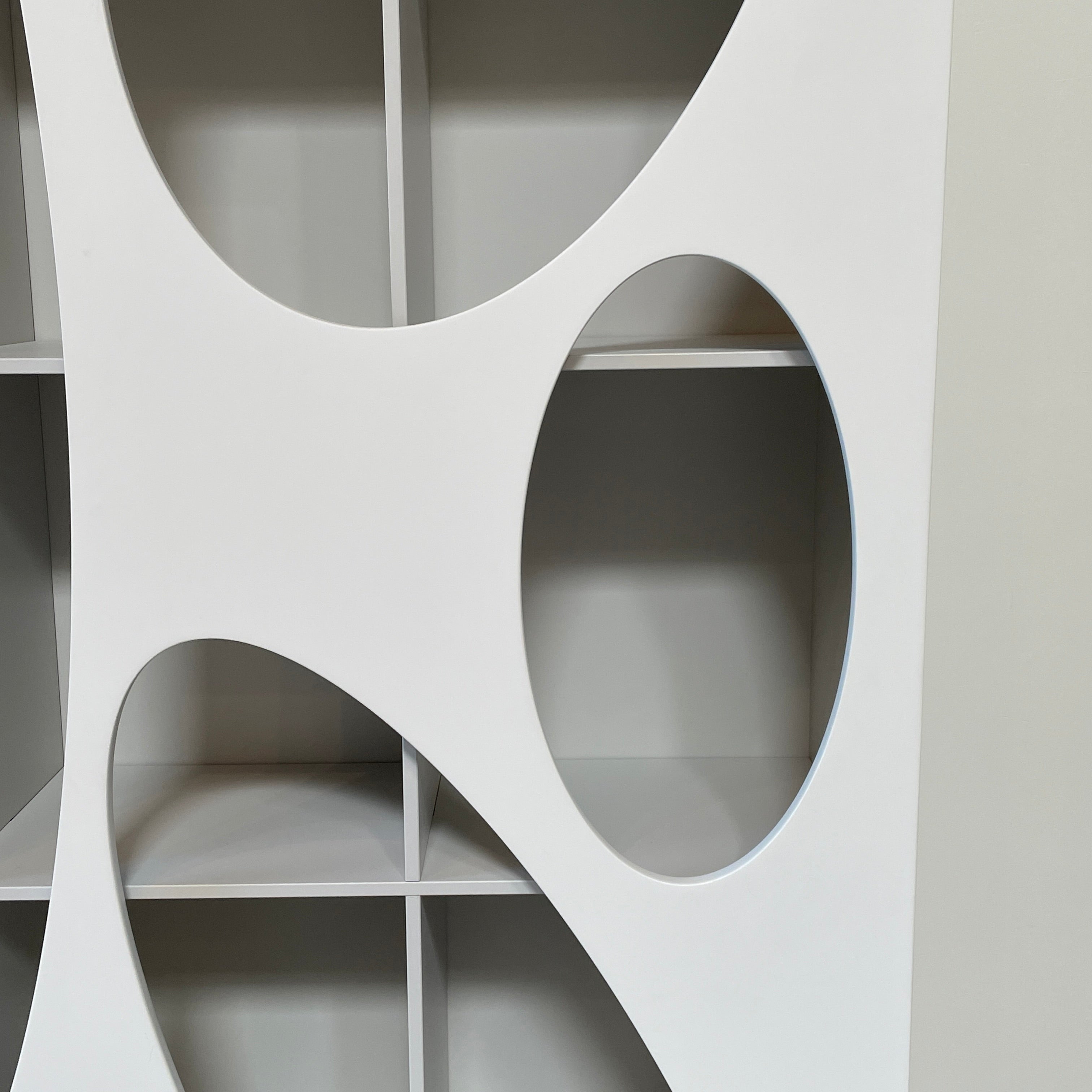 The Roche Bobois Denia Bookcase is a white bookshelf characterized by its abstract design, featuring multiple irregular oval cutouts, nine internal compartments, and integrated LED lighting, reminiscent of ROCHE BOBOIS styling.