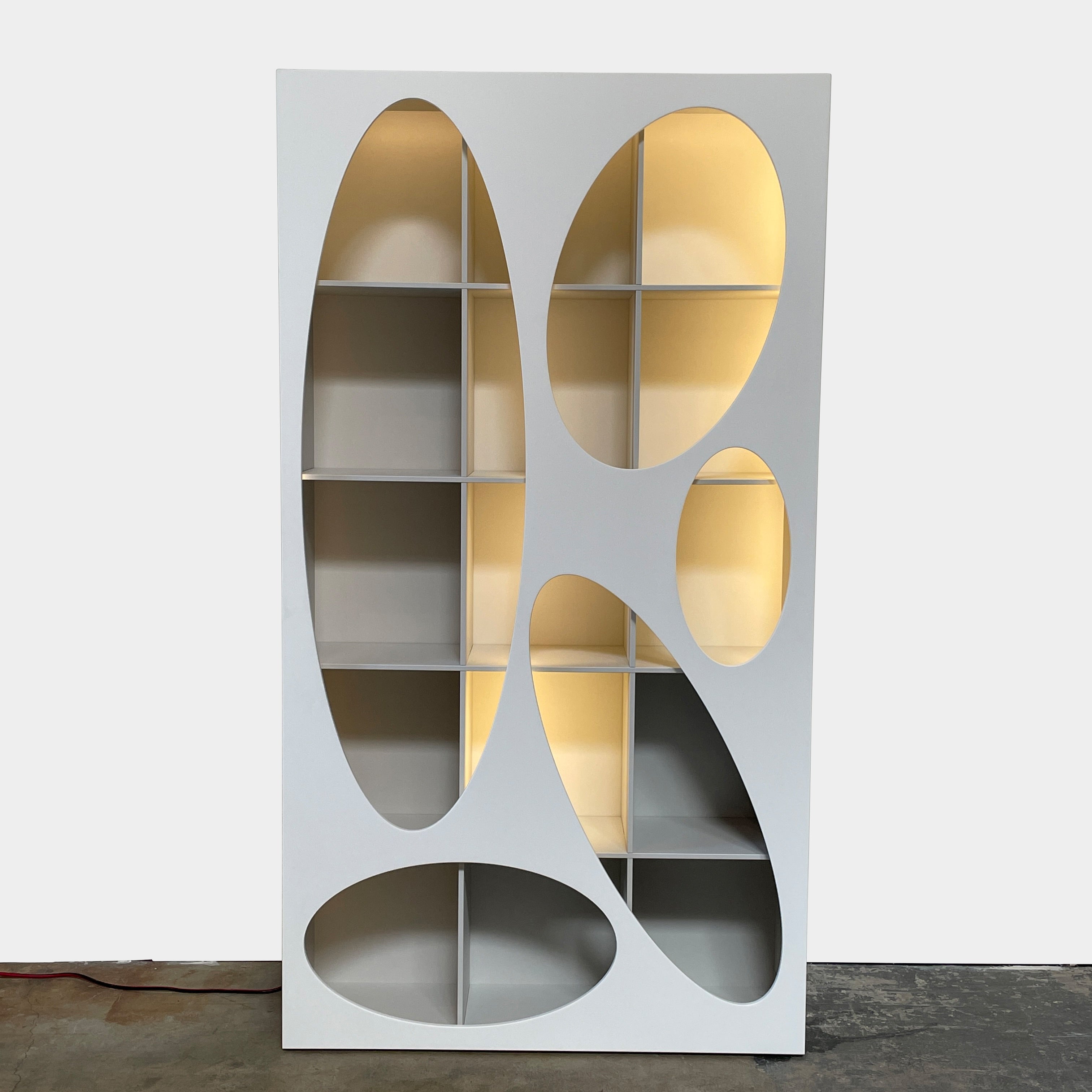 The Roche Bobois Denia Bookcase is a white bookshelf characterized by its abstract design, featuring multiple irregular oval cutouts, nine internal compartments, and integrated LED lighting, reminiscent of ROCHE BOBOIS styling.