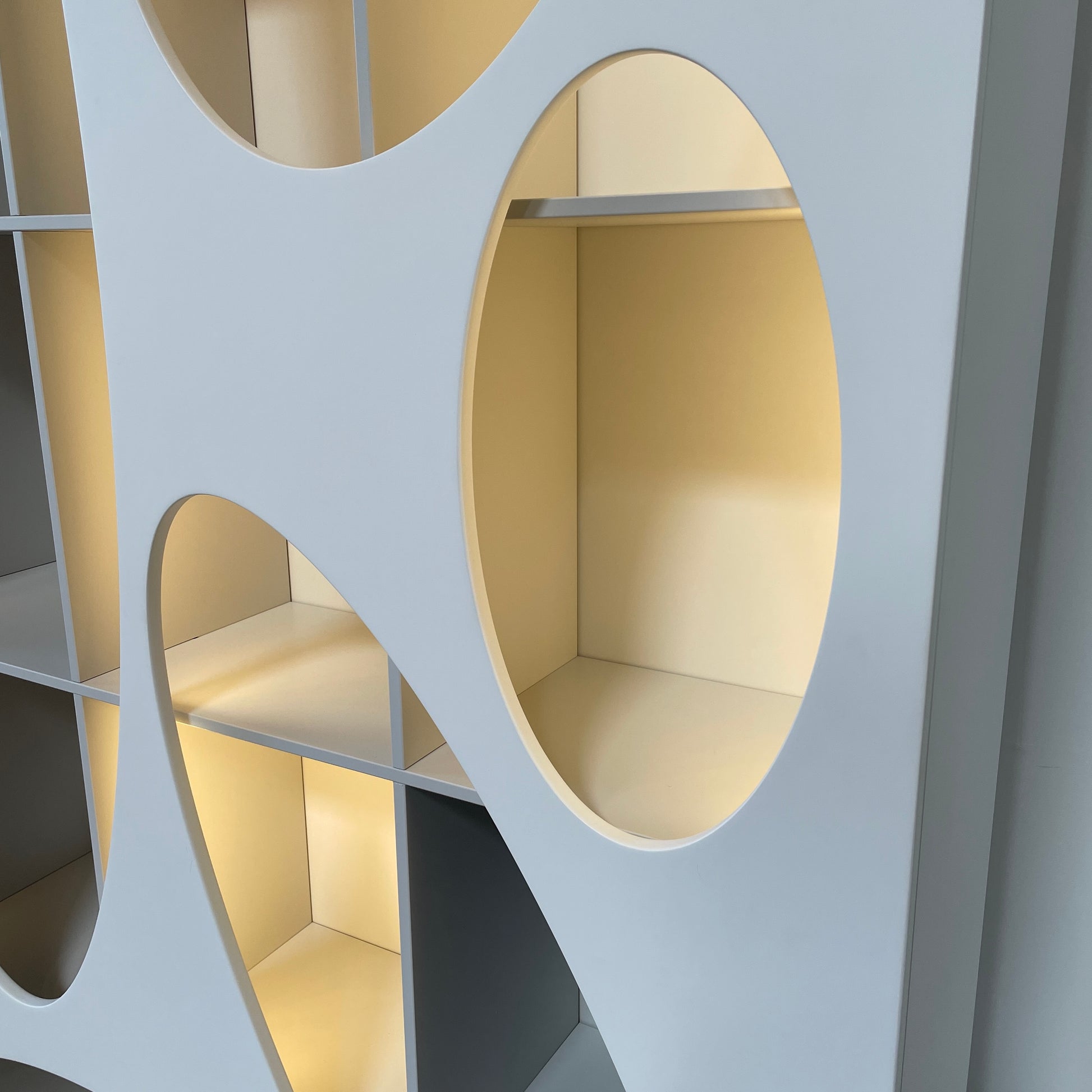 The Roche Bobois Denia Bookcase is a white bookshelf characterized by its abstract design, featuring multiple irregular oval cutouts, nine internal compartments, and integrated LED lighting, reminiscent of ROCHE BOBOIS styling.