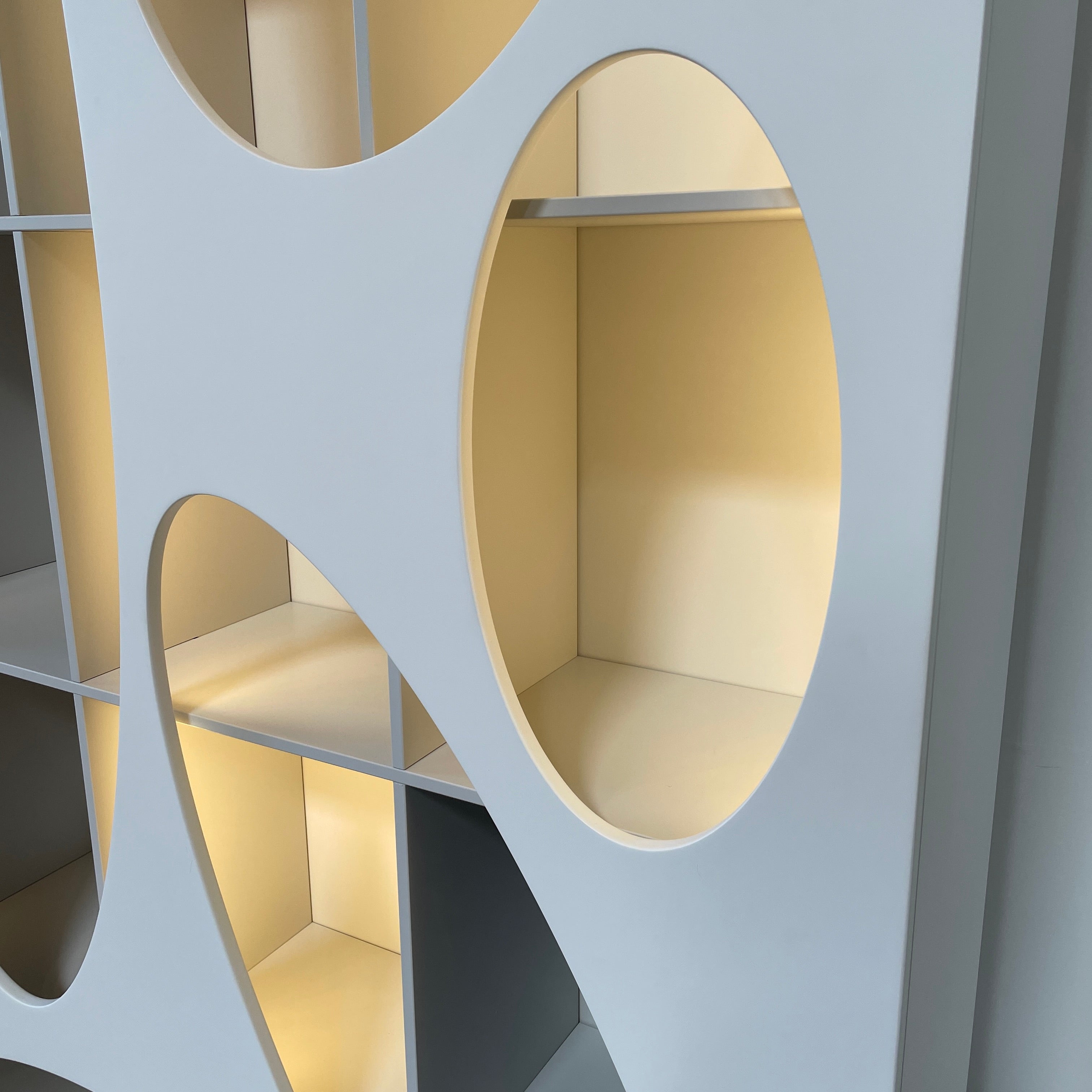 The Roche Bobois Denia Bookcase is a white bookshelf characterized by its abstract design, featuring multiple irregular oval cutouts, nine internal compartments, and integrated LED lighting, reminiscent of ROCHE BOBOIS styling.