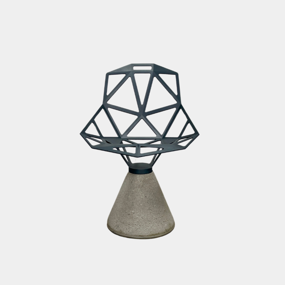 The Magis Grey Swivel Chair One With Concrete Base, designed by Konstantin Grcic, showcases a geometric open lattice design on a concrete base, sitting gracefully on a textured floor against a plain wall.