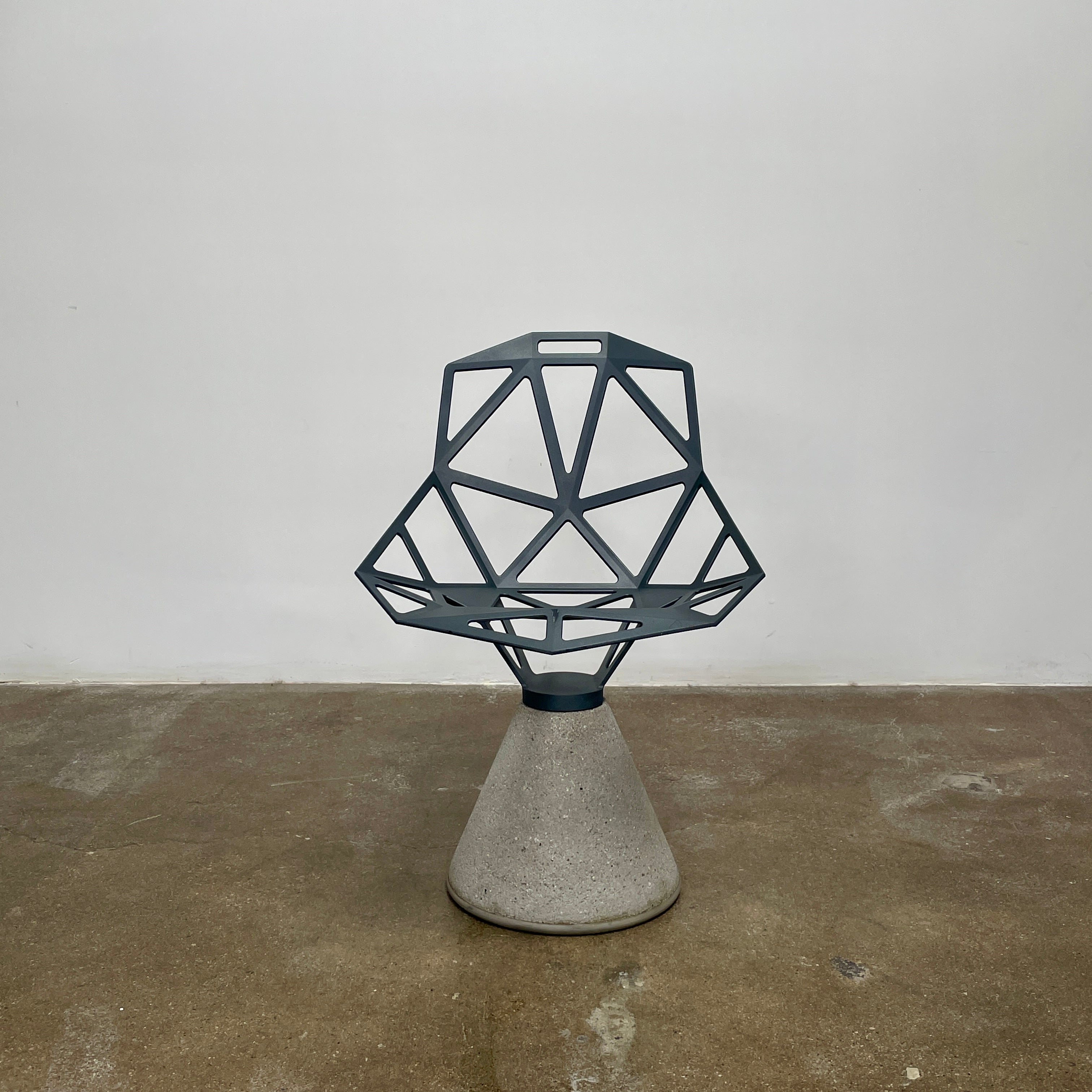 The Magis Grey Swivel Chair One With Concrete Base, designed by Konstantin Grcic, showcases a geometric open lattice design on a concrete base, sitting gracefully on a textured floor against a plain wall.