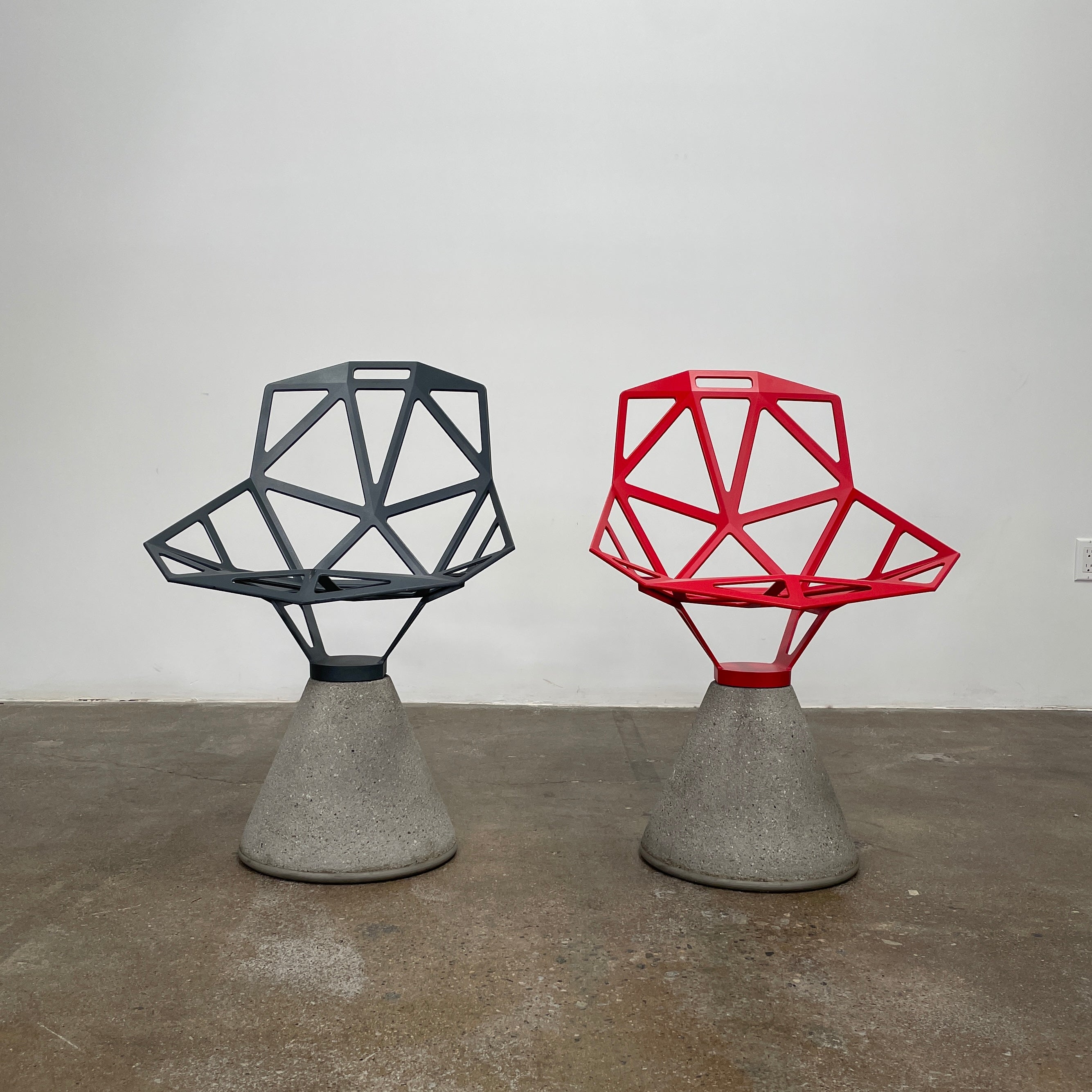 The Magis Grey Swivel Chair One With Concrete Base, designed by Konstantin Grcic, showcases a geometric open lattice design on a concrete base, sitting gracefully on a textured floor against a plain wall.