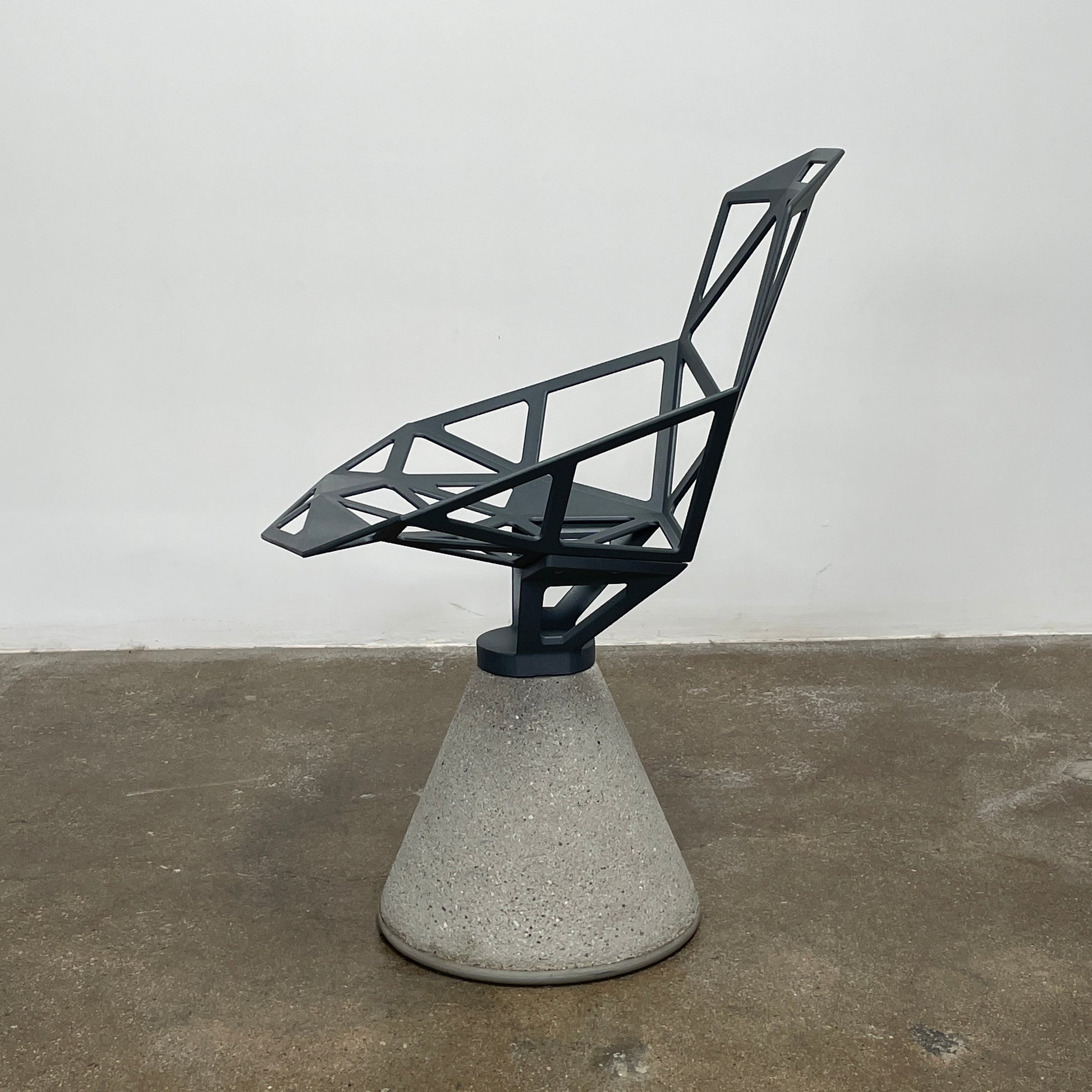 The Magis Grey Swivel Chair One With Concrete Base, designed by Konstantin Grcic, showcases a geometric open lattice design on a concrete base, sitting gracefully on a textured floor against a plain wall.